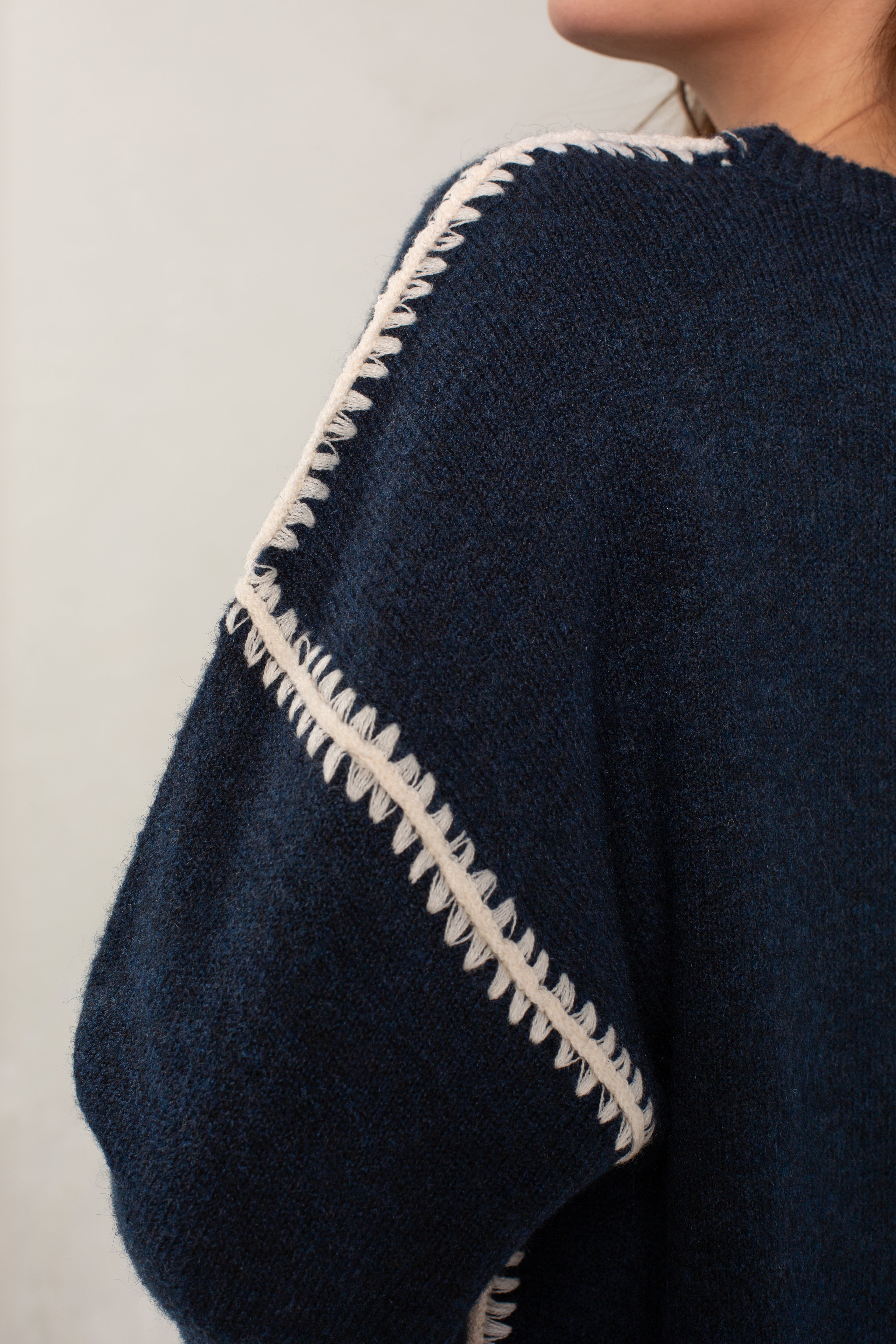 Close-up of a person wearing Isla's The Jackie Sweater—an oversized, dark blue textured knit with light blanket stitching along the shoulder and sleeve seam. Only the upper body and shoulder are visible.