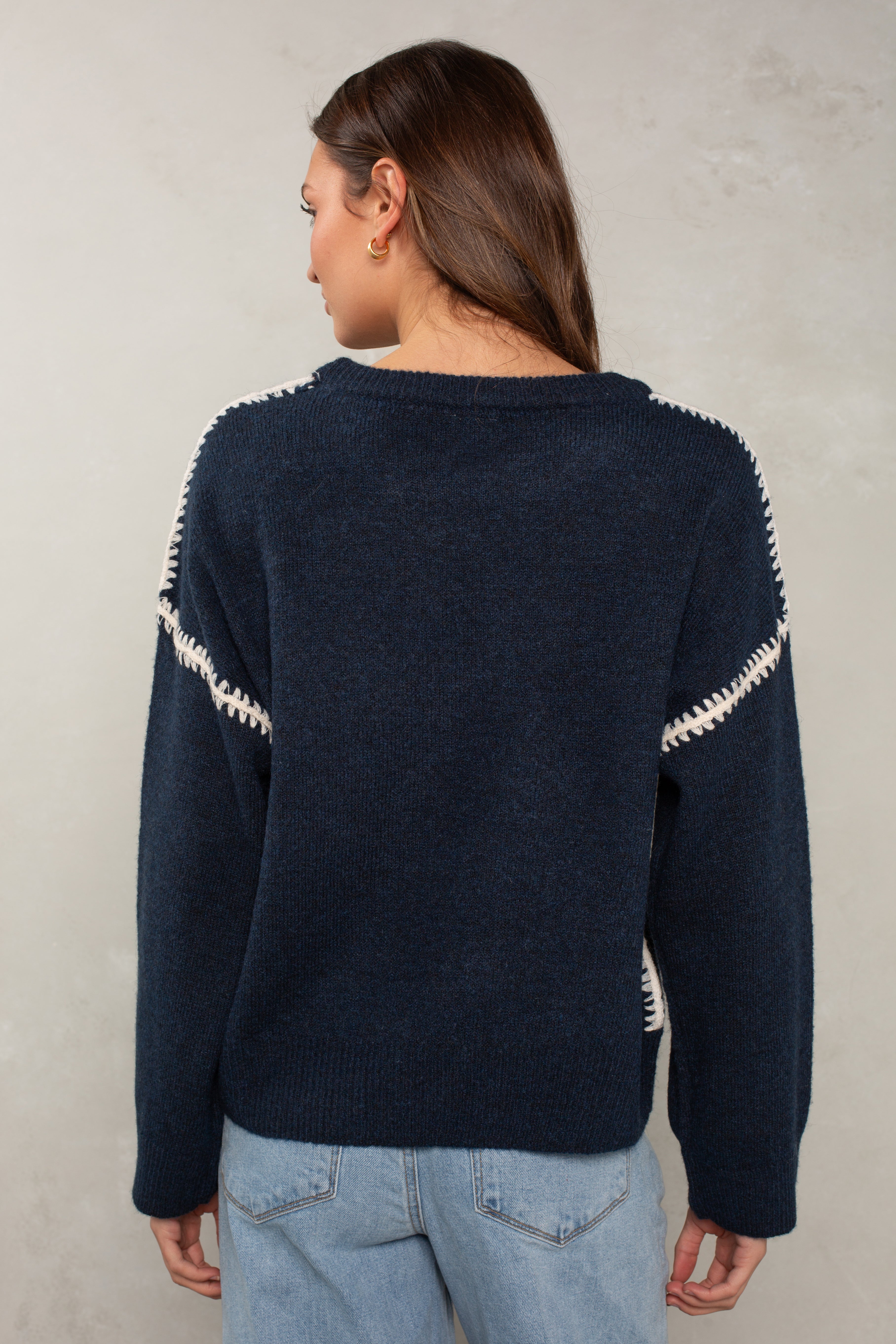 A woman with long brown hair, wearing Isla’s The Jackie Sweater in navy blue with white zigzag and blanket stitching on the shoulders and light blue jeans, stands facing away against a neutral beige background.
