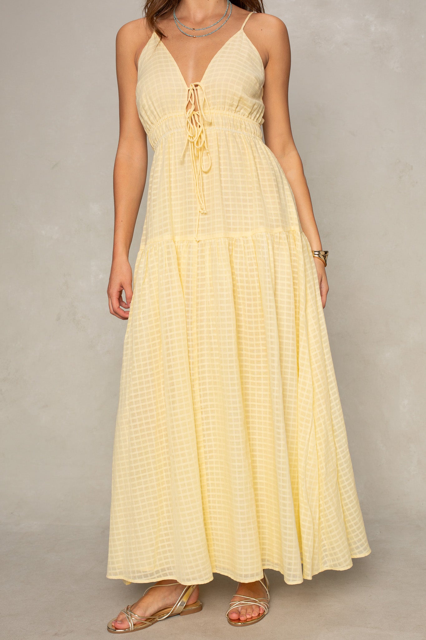 A woman wears the Isla Kallie Tiered Maxi Dress in light yellow, featuring a deep V-neckline, front tie, and flowy skirt. She stands against a neutral background, styled with strappy sandals and jewelry.