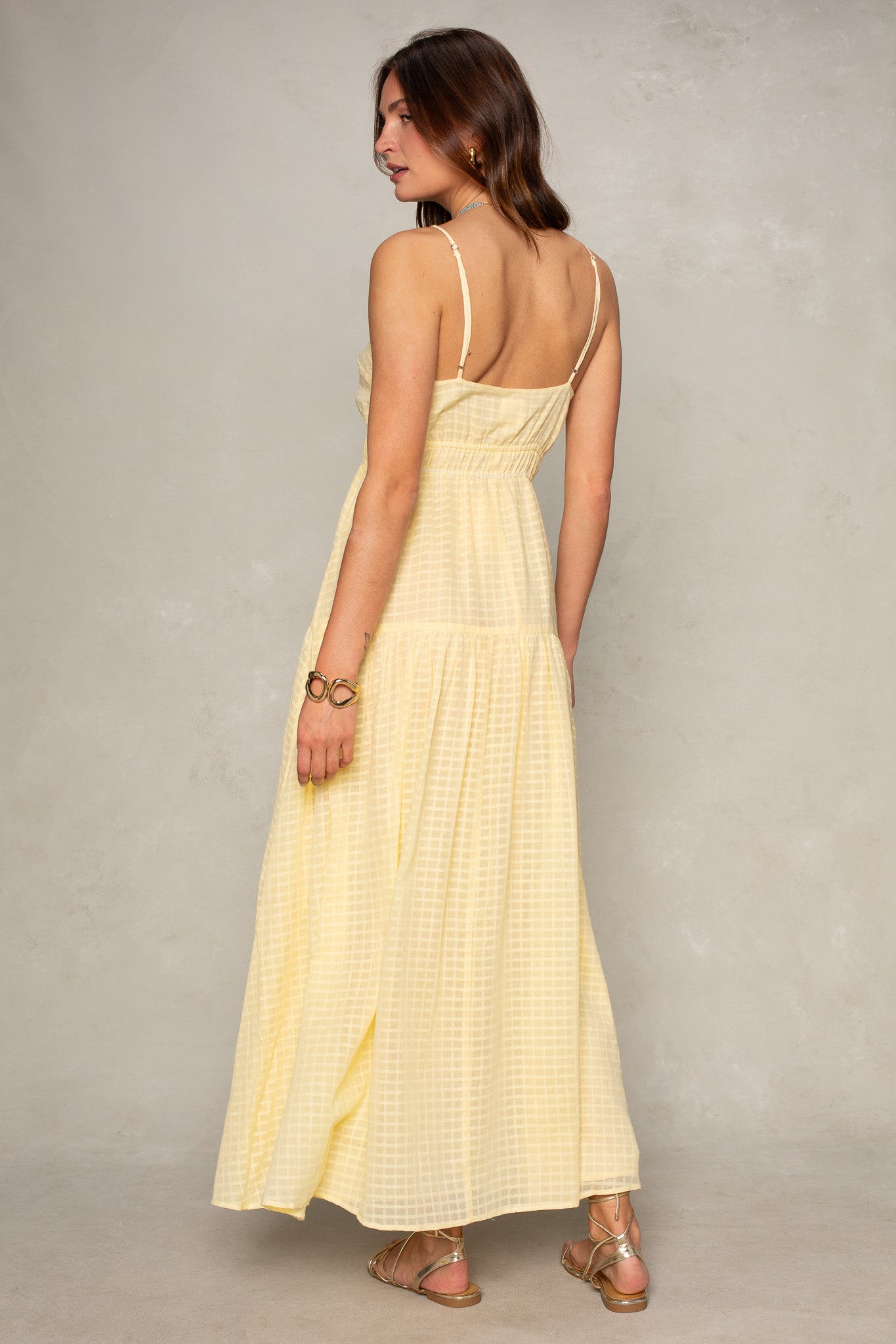 A woman models the Isla Kallie Tiered Maxi Dress—a light yellow, sleeveless, ankle-length dress with thin straps and a subtle checked pattern—against a neutral background. She is turned slightly away, showcasing the back.