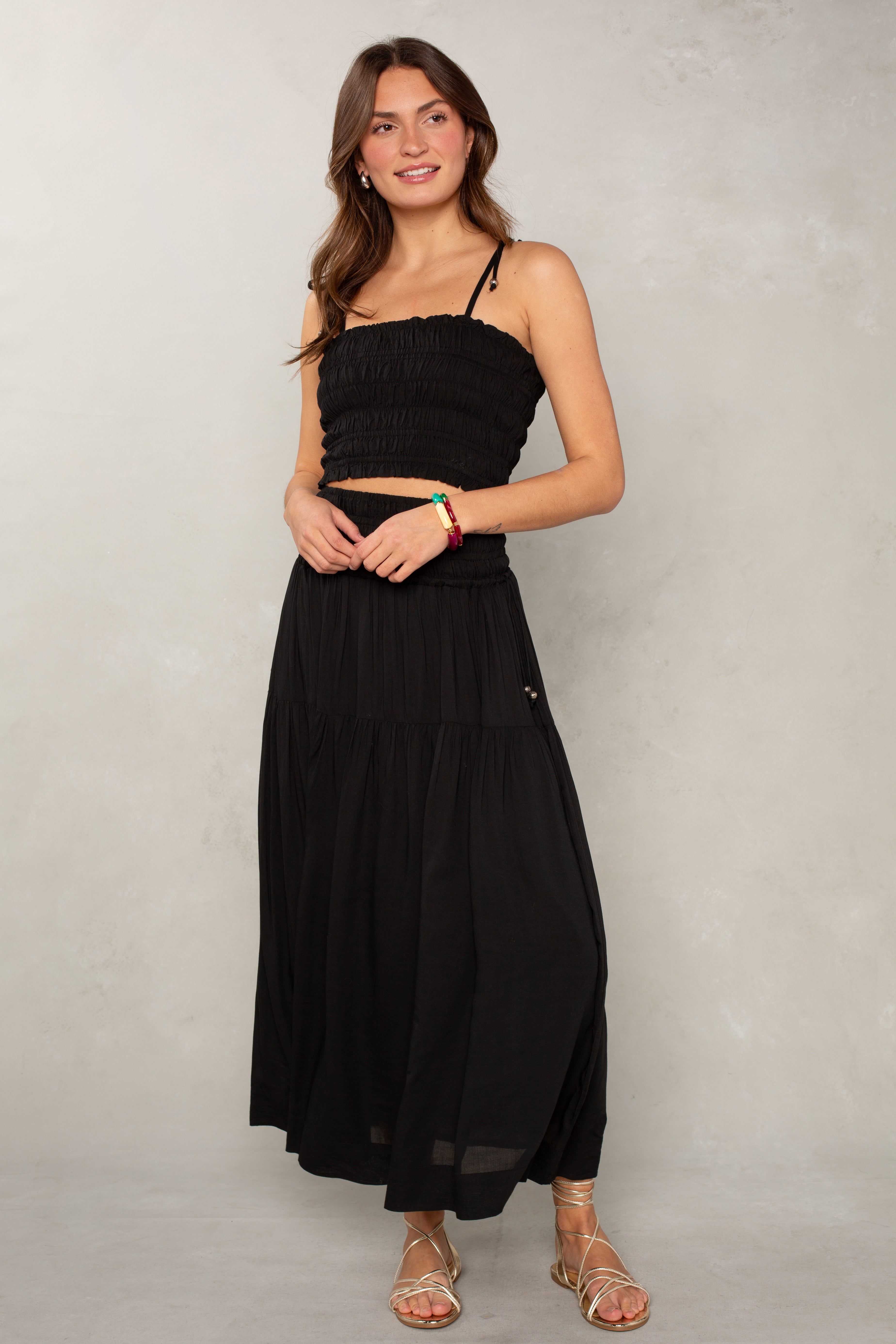 A woman wearing the Isla Jules Smocked Skirt in Black paired with a black sleeveless crop top, gold sandals, and colorful bracelets stands against a light gray textured wall—a chic take on effortless summer style.