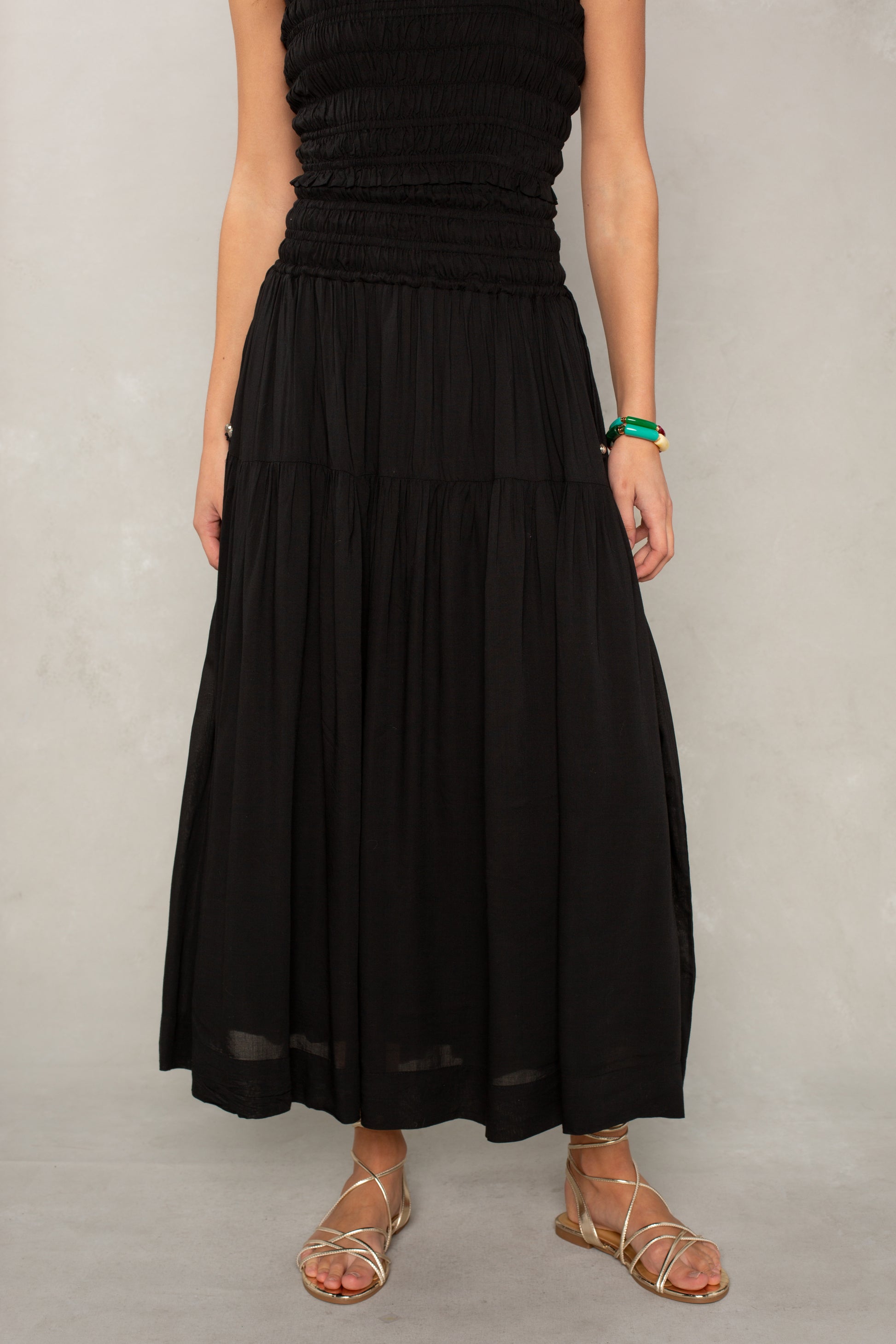 A person stands against a light gray background wearing the Jules Smocked Skirt in Black by Isla, styled with gold sandals. The skirt features a smocked waistband and flows gracefully while their arms rest by their sides, and their face is not visible in the image.