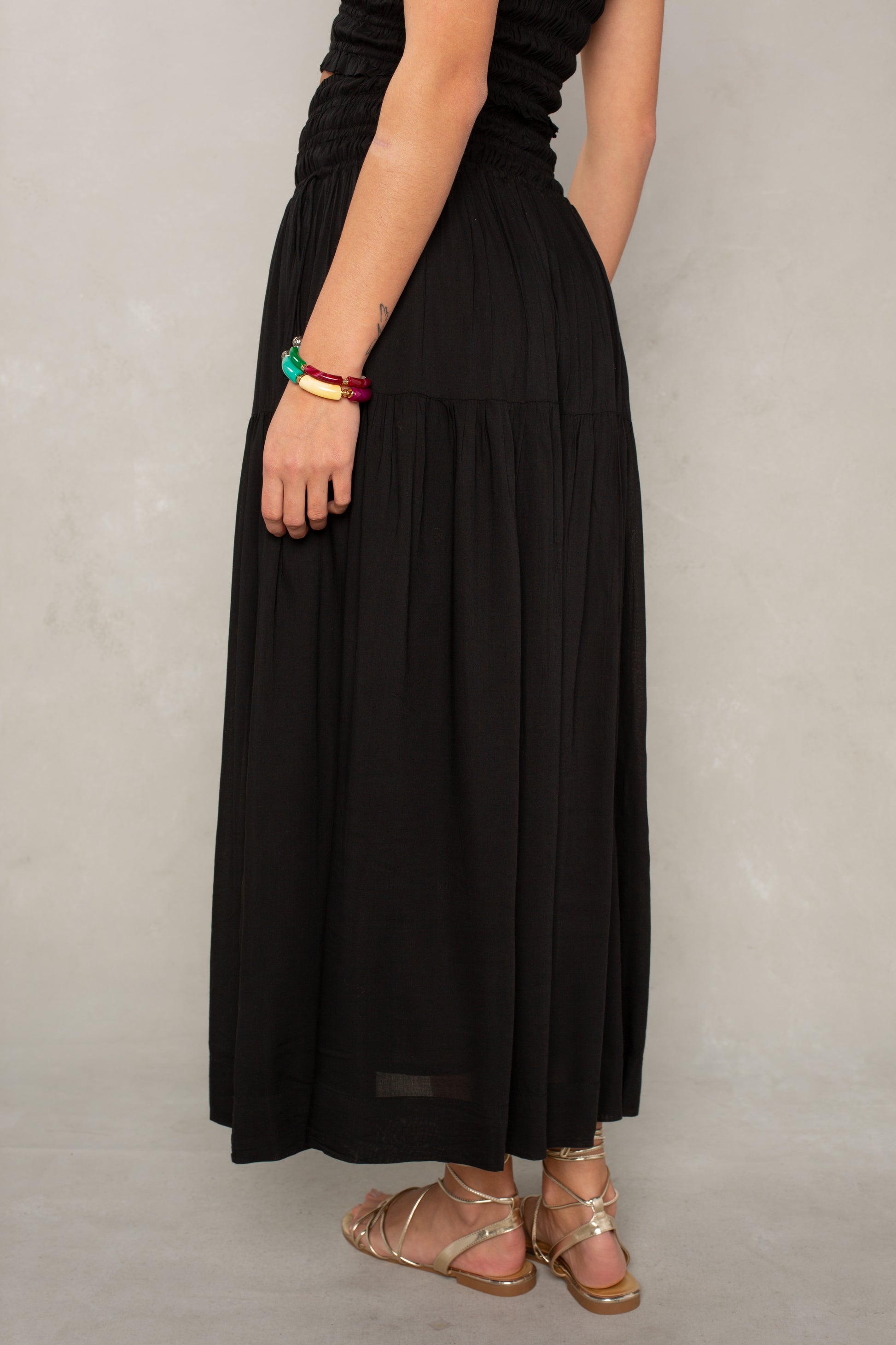 A person stands against a light gray background, wearing the Isla Jules Smocked Skirt in Black, paired with a black top and sandals. Only the lower half and one arm are visible, accessorized with two chunky bracelets. The skirt is made from breathable fabric.