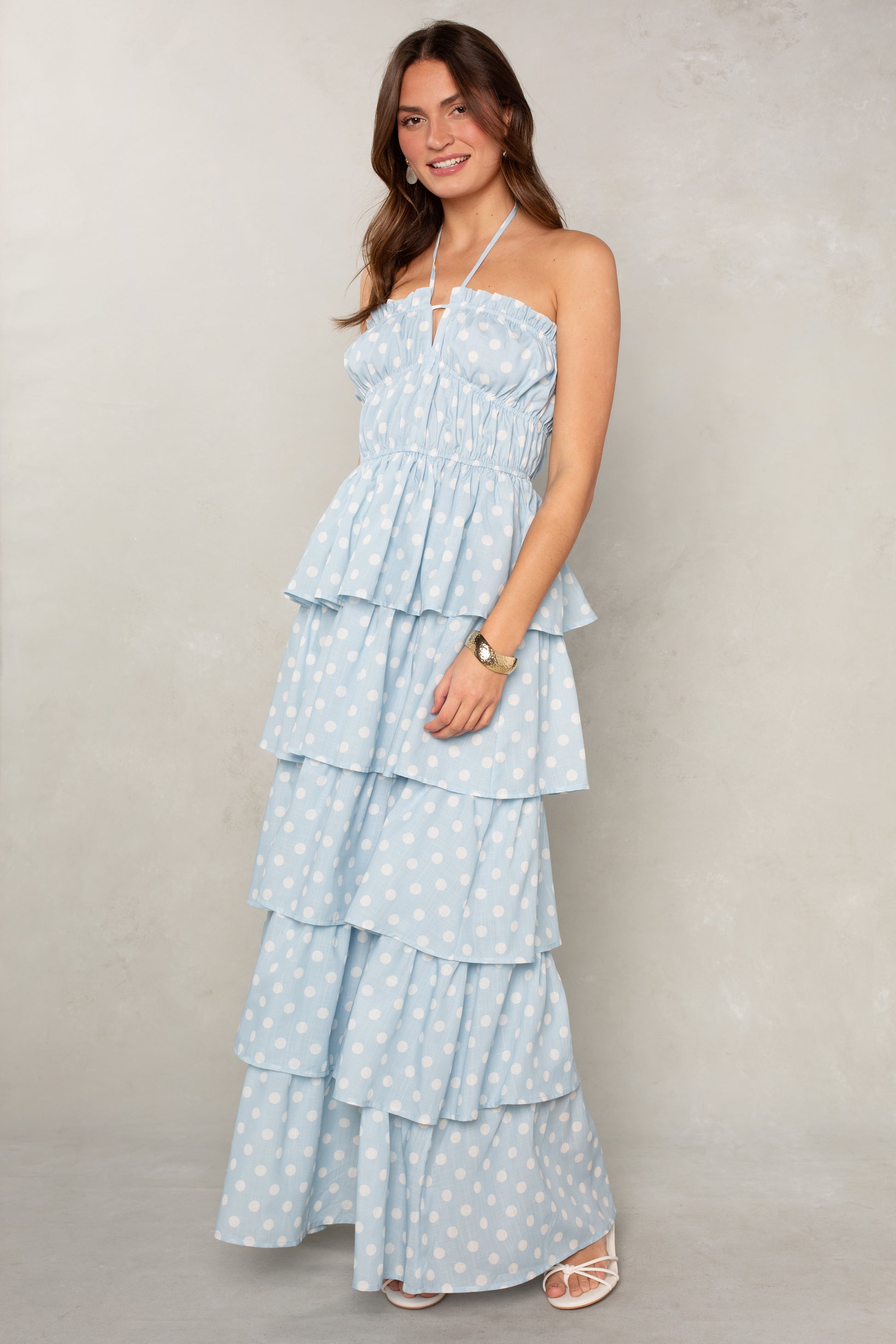 A woman smiles in the Isla Lindsey Ruffle Tiered Maxi, a light blue halter dress with cascading ruffles and white polka dots, paired with white sandals. She has long brown hair and poses against a neutral light background.