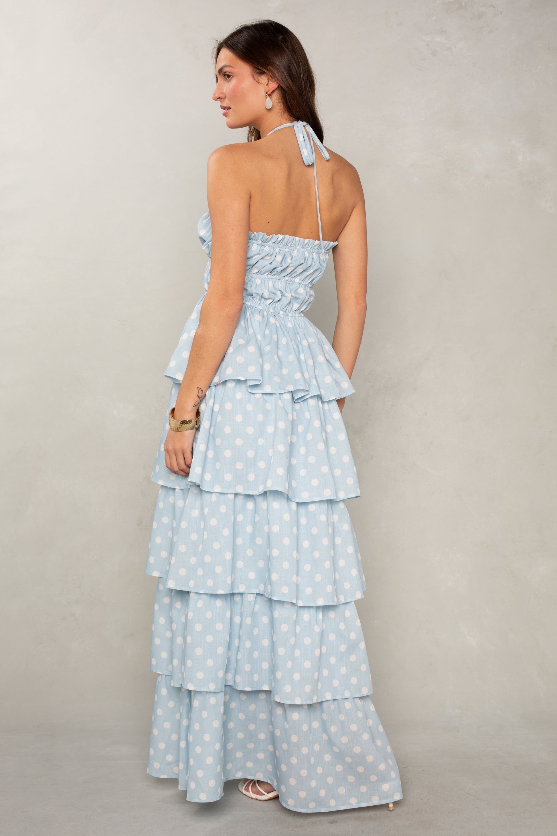 A woman with long brown hair wears the Isla Lindsey Ruffle Tiered Maxi, a light blue halter-neck dress with white polka dots and cascading ruffles, paired with hoop earrings and white sandals against a light gray background.
