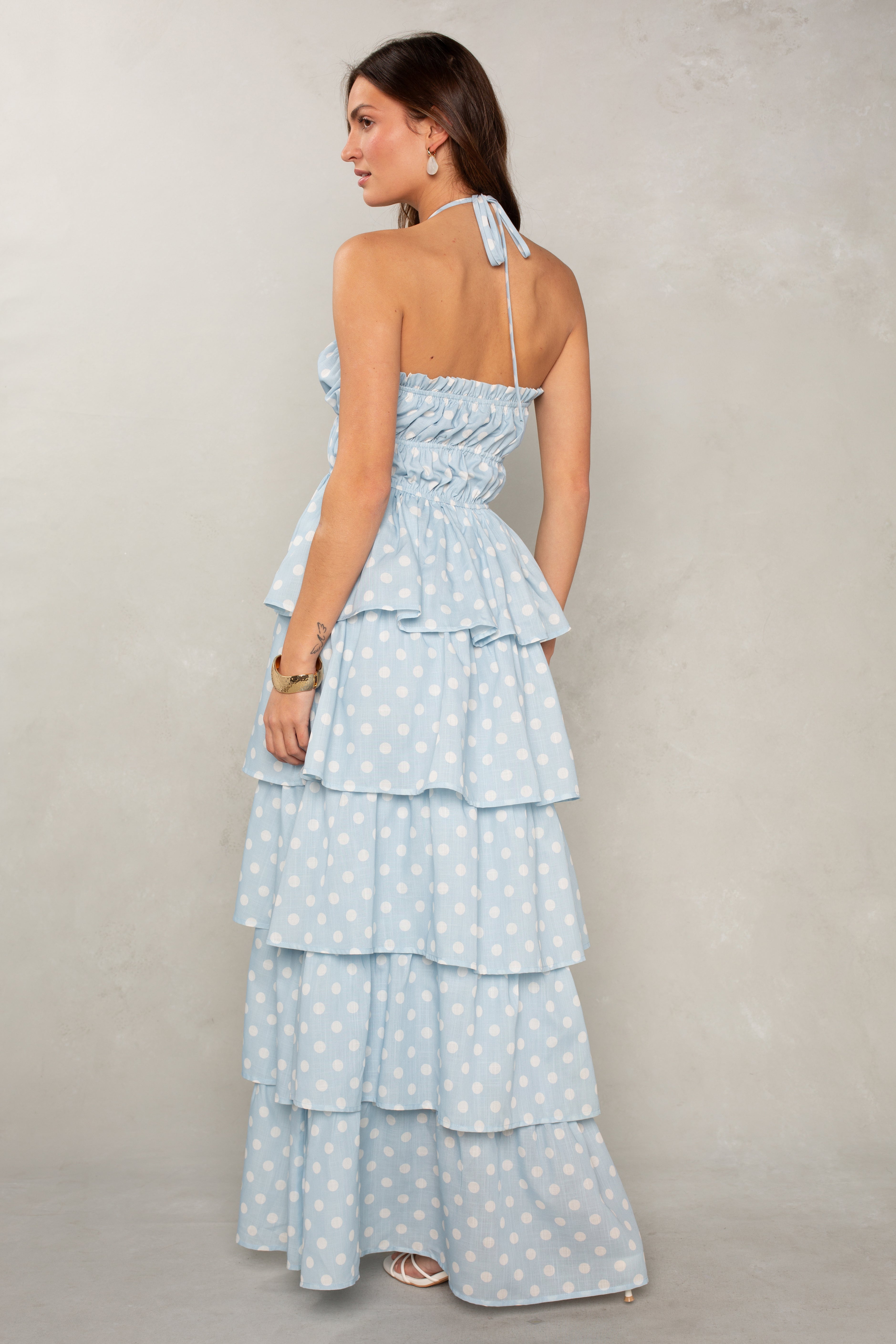 A woman with long brown hair wears the Isla Lindsey Ruffle Tiered Maxi, a light blue halter-neck dress with white polka dots and cascading ruffles, paired with hoop earrings and white sandals against a light gray background.
