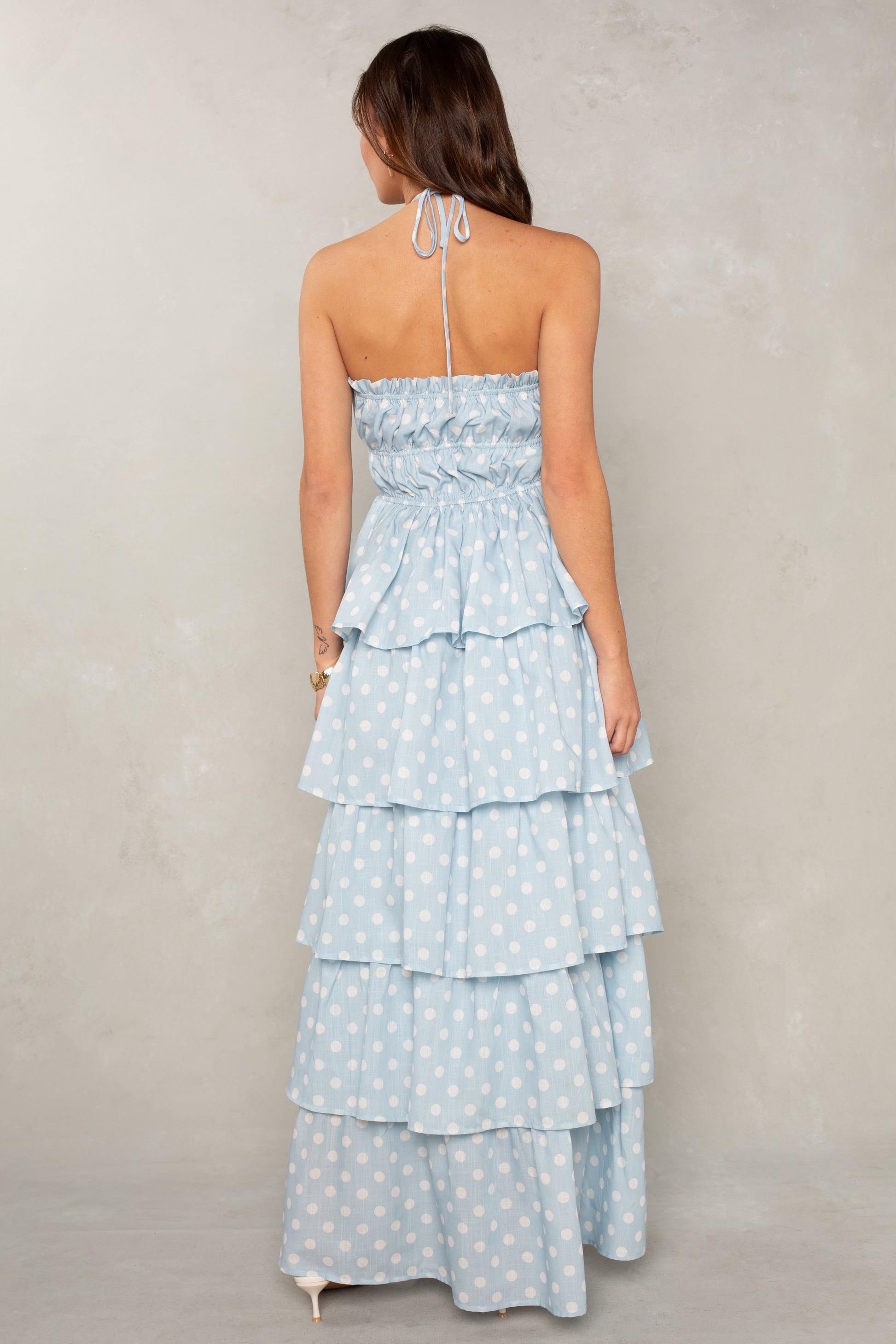 Woman facing away in the Isla Lindsey Ruffle Tiered Maxi—a blue, sleeveless, halter-neck dress with white polka dots and cascading ruffles. Her hair is down as she stands against a neutral background.