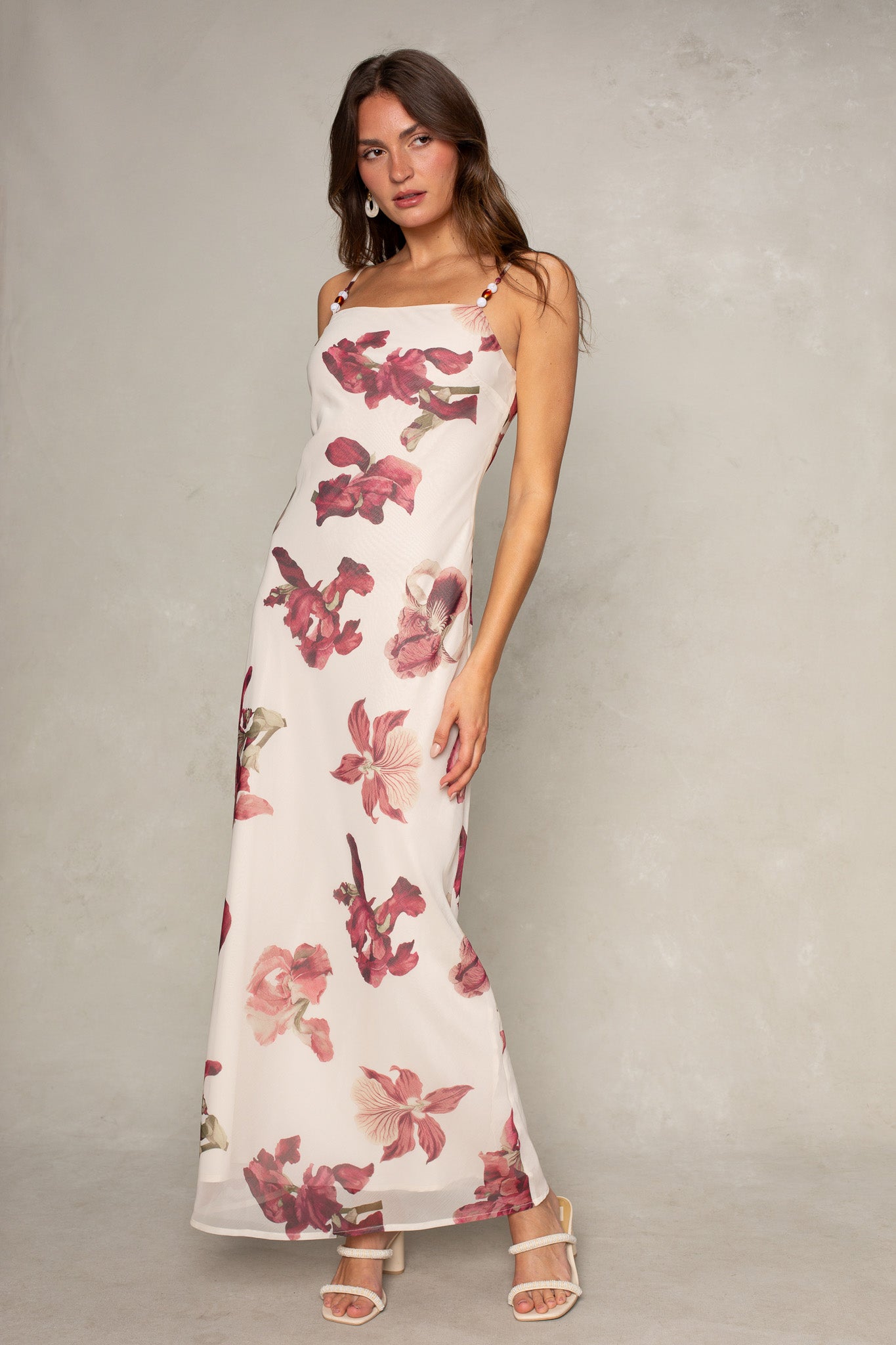 A woman poses with one hand on her hip against a neutral background, wearing the Isla Isa Beaded Strap Column Maxi Dress. She has long brown hair and white sandals, showcasing this sleeveless, floor-length dress ideal for formal occasions.