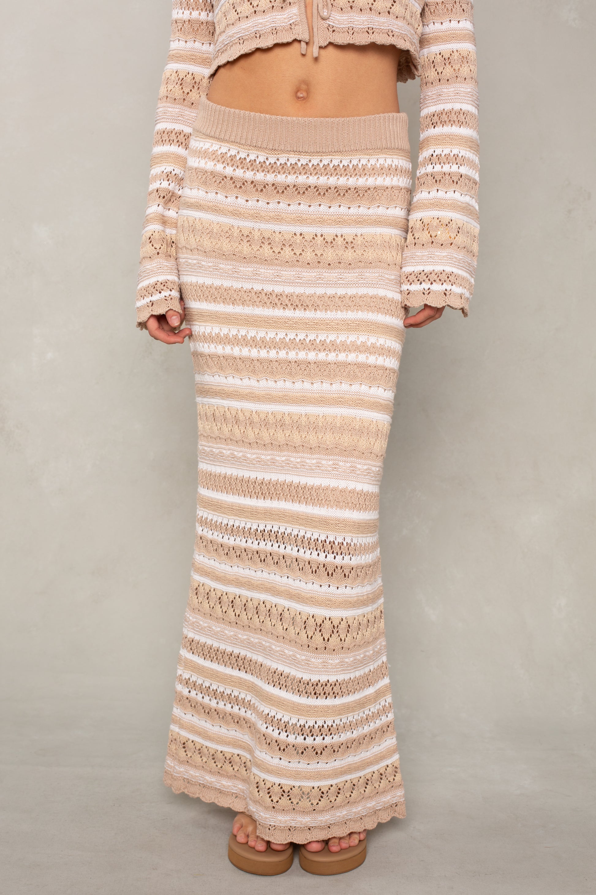 A person wears the Isla Andrea Maxi Skirt, a high-waisted cream and beige crochet knit with horizontal stripes, paired with its matching long-sleeve crop top and sandals against a neutral background.