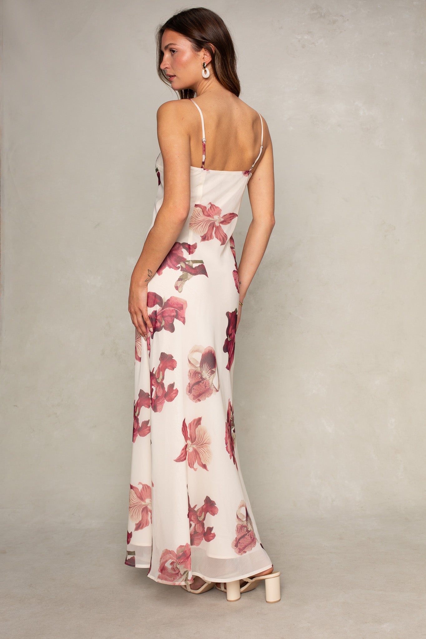 A woman faces away in the Isla Isa Beaded Strap Column Maxi Dress, featuring pink and red floral prints. She has long brown hair and pairs the formal look with platform heels and statement earrings against a simple light background.