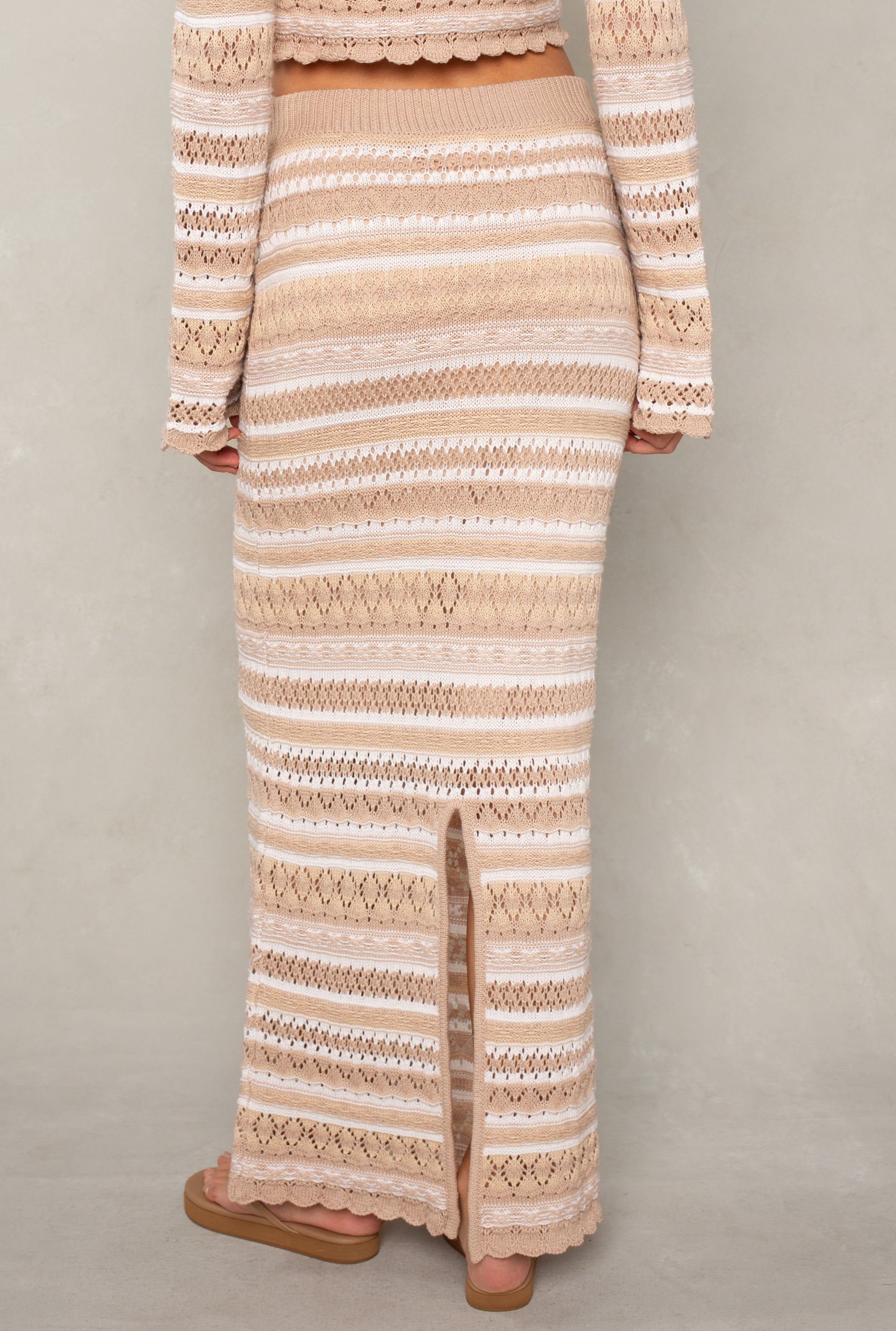A person wearing the Isla Andrea Maxi Skirt—a high-waisted, beige knit skirt with white stripes and a back slit—paired with a matching top and tan sandals, stands against a light gray background.