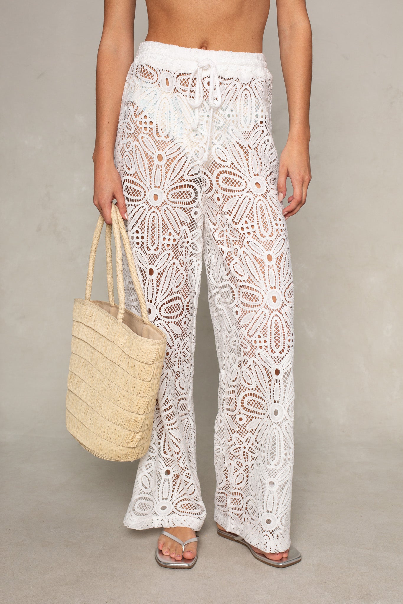A person wears the Isla Maisie Lace Coverup Pant in white, paired with a woven straw tote and flat sandals—ideal for a resort-ready, warm-weather look. The background is plain and neutral.