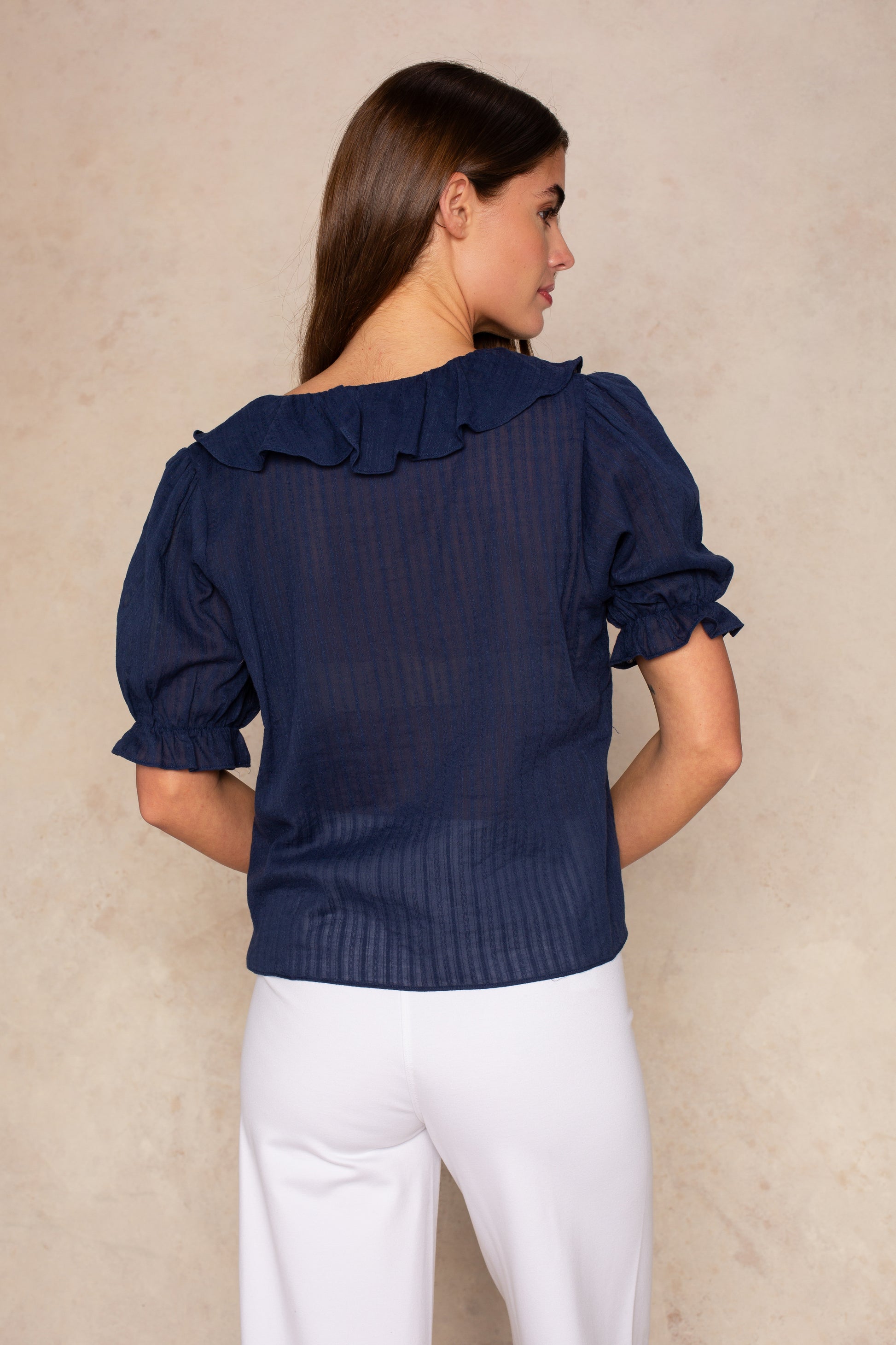 A woman with long brown hair stands facing away, wearing the Georgie Aimee Ruffle Short Sleeve Blouse in navy with white pants, made of lightweight textured fabric, against a beige textured background.