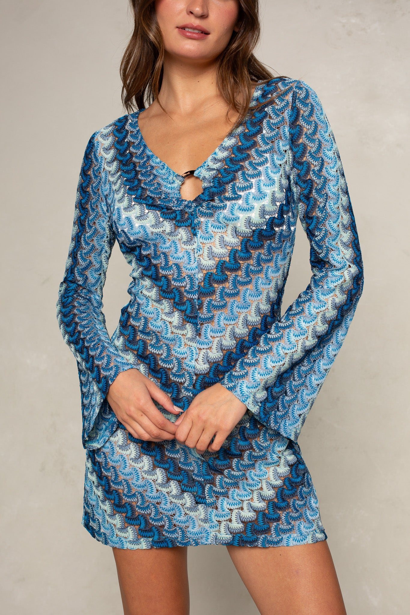 A woman stands against a light background, wearing the Isla Stella O-ring Mini Coverup—a blue and white long-sleeve beach dress with an o-ring neckline and front keyhole. Her face is partially visible.