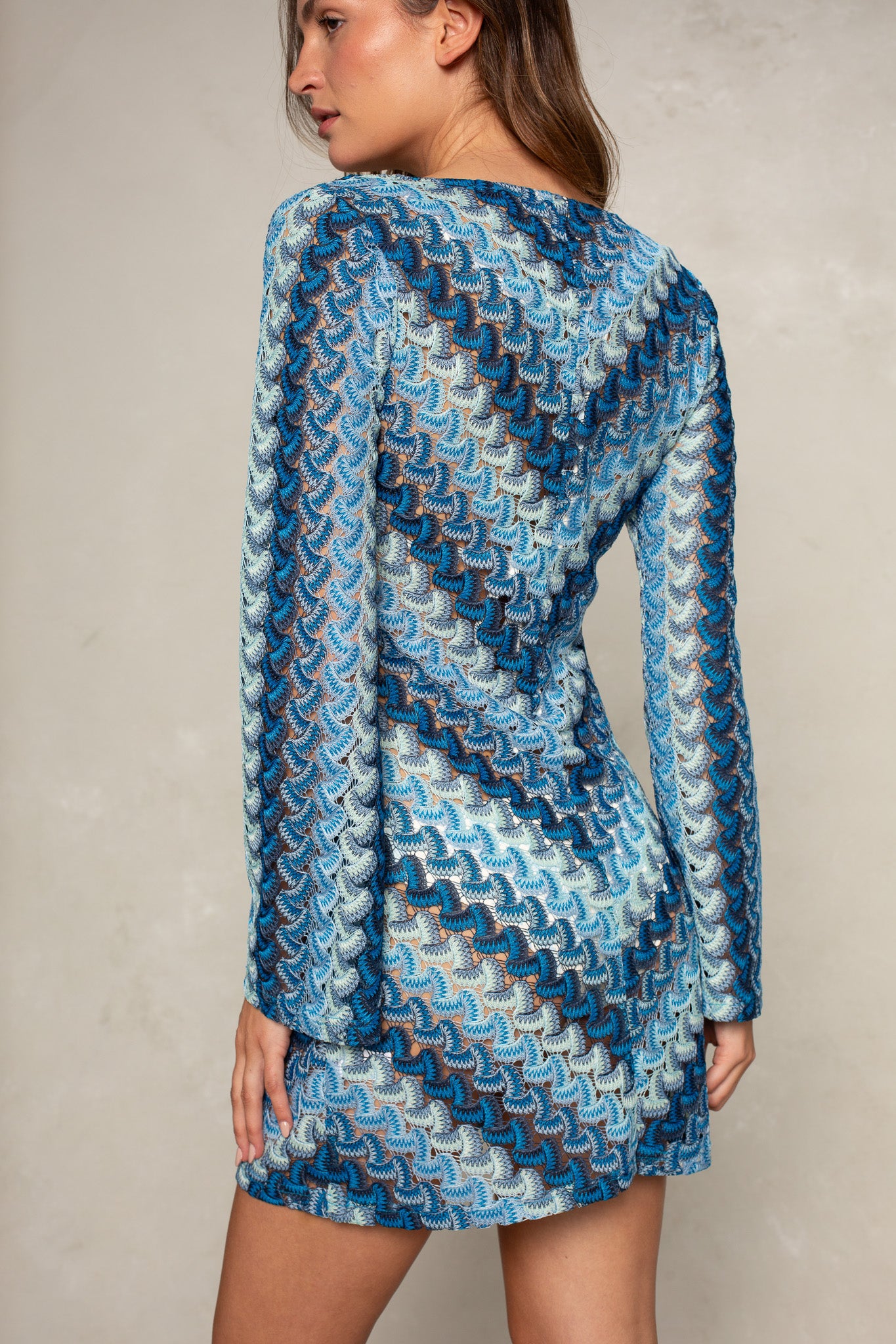 A woman poses from behind against a neutral background in the Isla Stella O-ring Mini Coverup, a long-sleeve, fitted beach dress featuring a blue, turquoise, and white zigzag-patterned knit design.