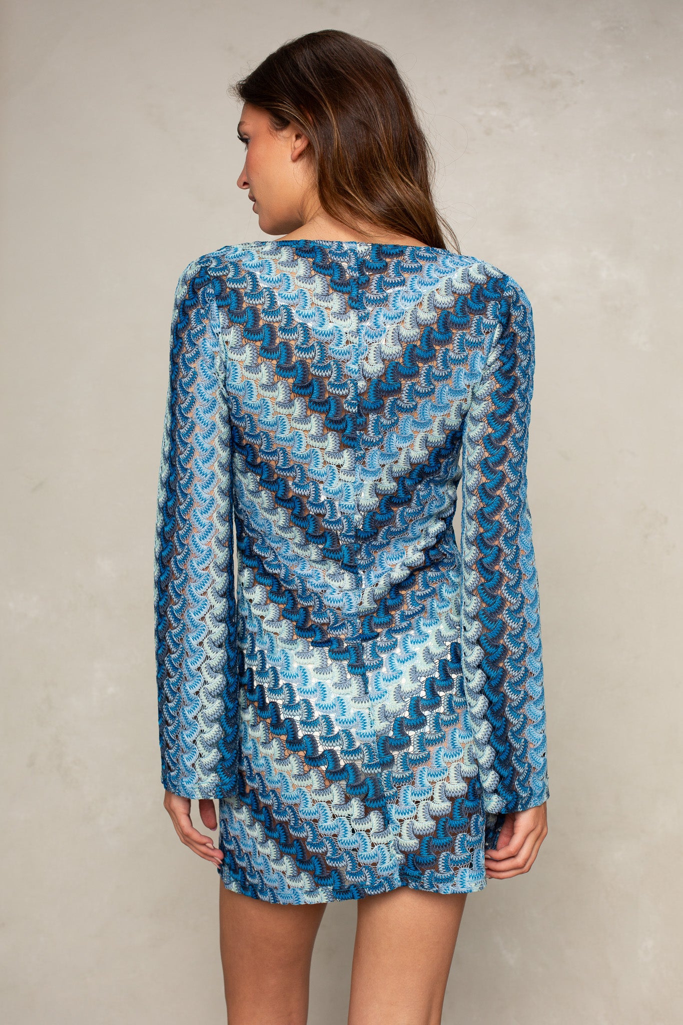 A person with long brown hair faces away, wearing the Isla Stella O-ring Mini Coverup—a blue, long-sleeve mini dress with a textured zigzag pattern—against a plain, light-colored wall.