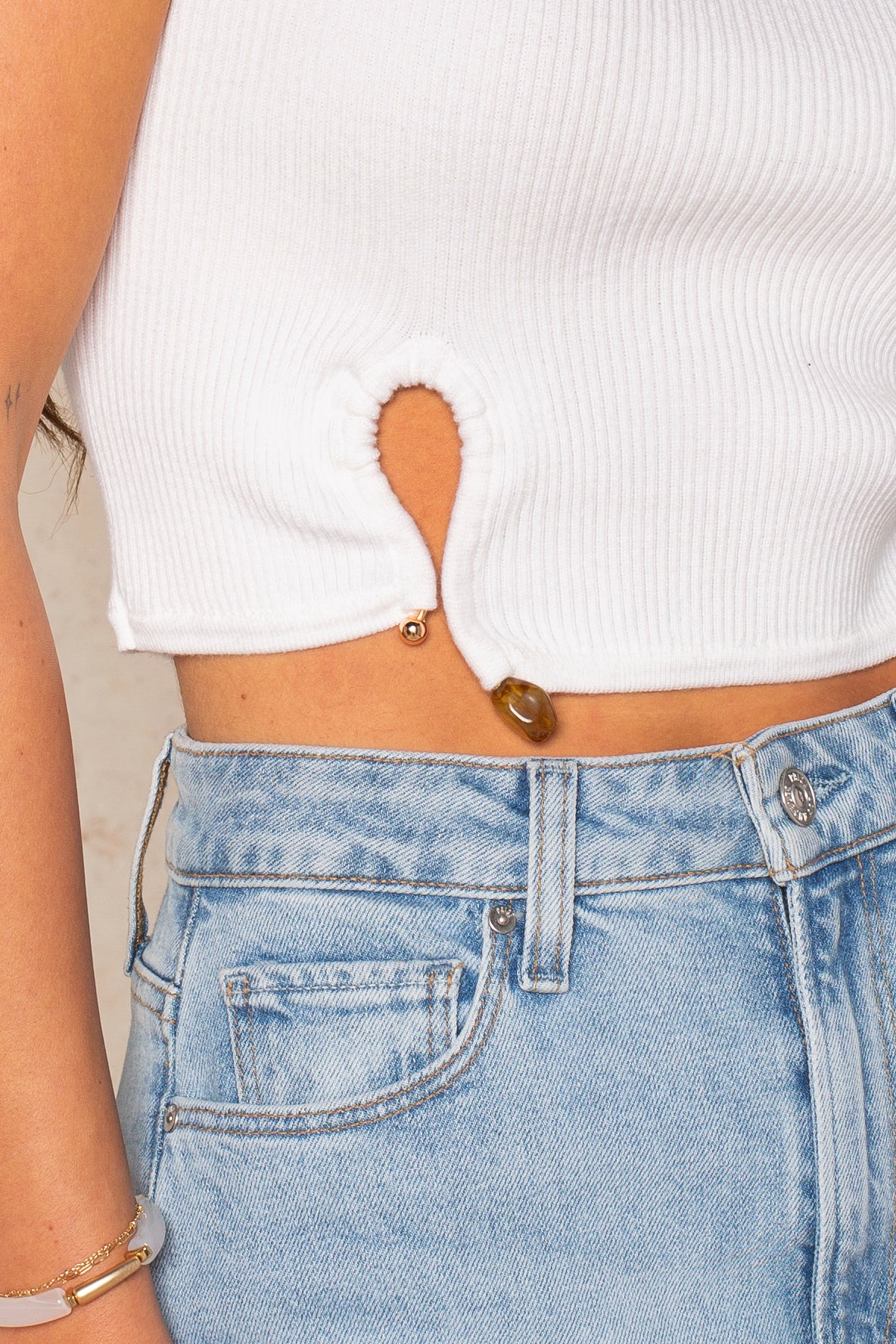 A close-up of a person wearing Isla’s Tyler Hardware Sweater Tank—a white ribbed cropped top with a small keyhole cutout above the hem—paired with blue high-waisted jeans and a visible belly button piercing.