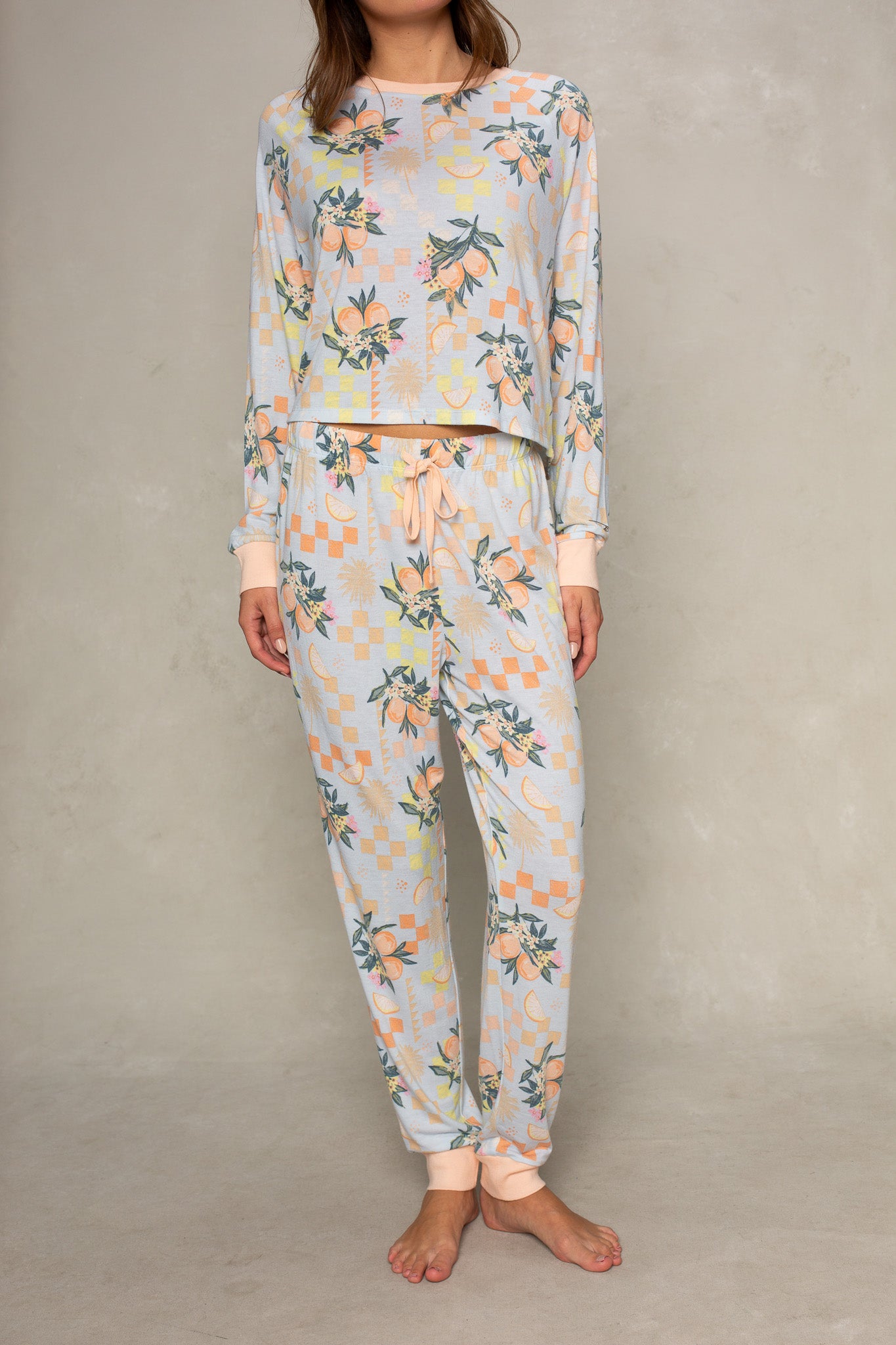 A woman stands barefoot in the Isla Citrus Check Brushed Jersey Pant in Orange Multi, paired with a matching relaxed-fit long-sleeve top, both featuring a floral and geometric pattern, against a light neutral background.