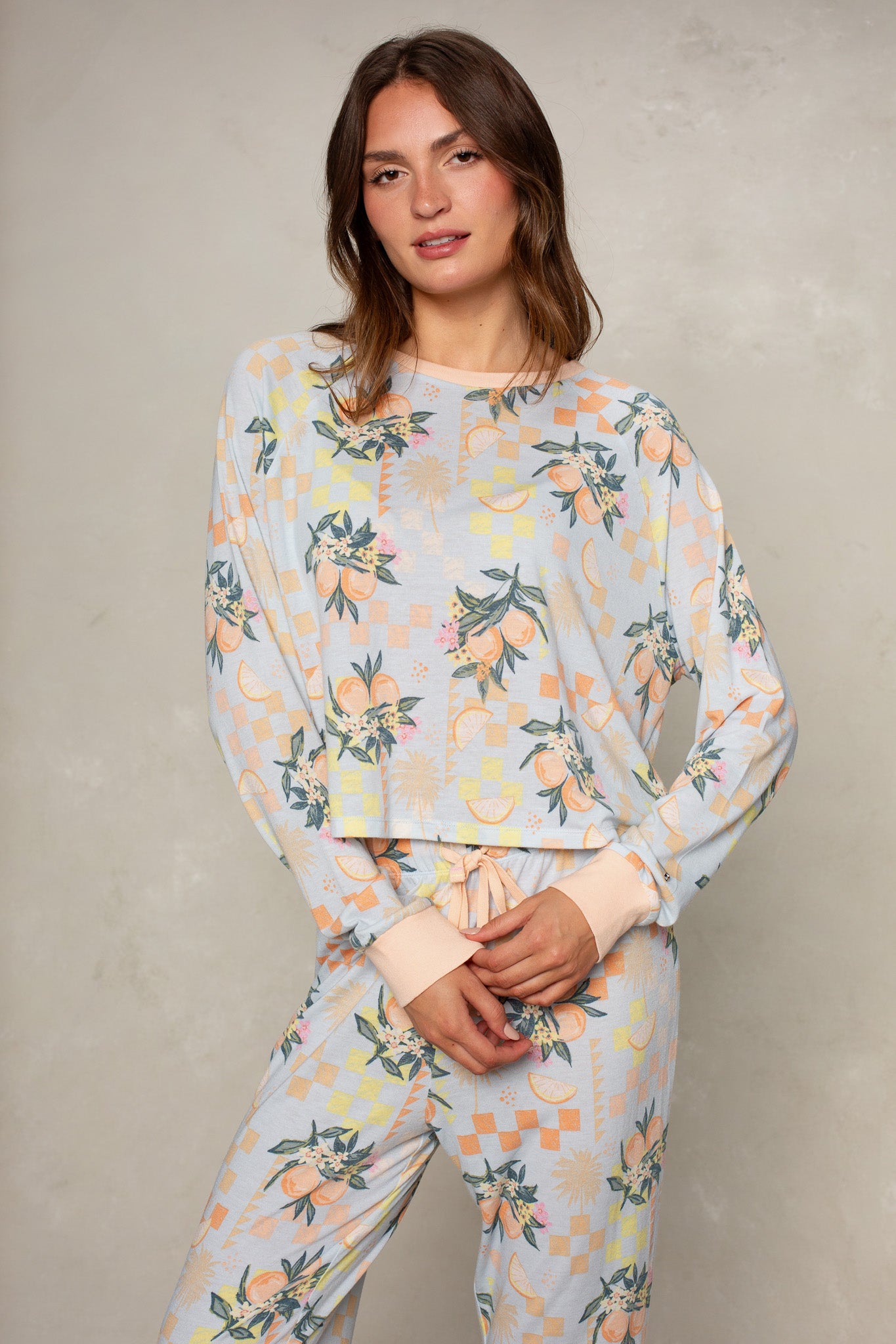 A woman with long brown hair models the Isla Citrus Check Brushed Jersey Top in Orange Multi, featuring a relaxed fit and geometric-floral pattern. She stands against a light grey background, looking at the camera—ideal for layering.