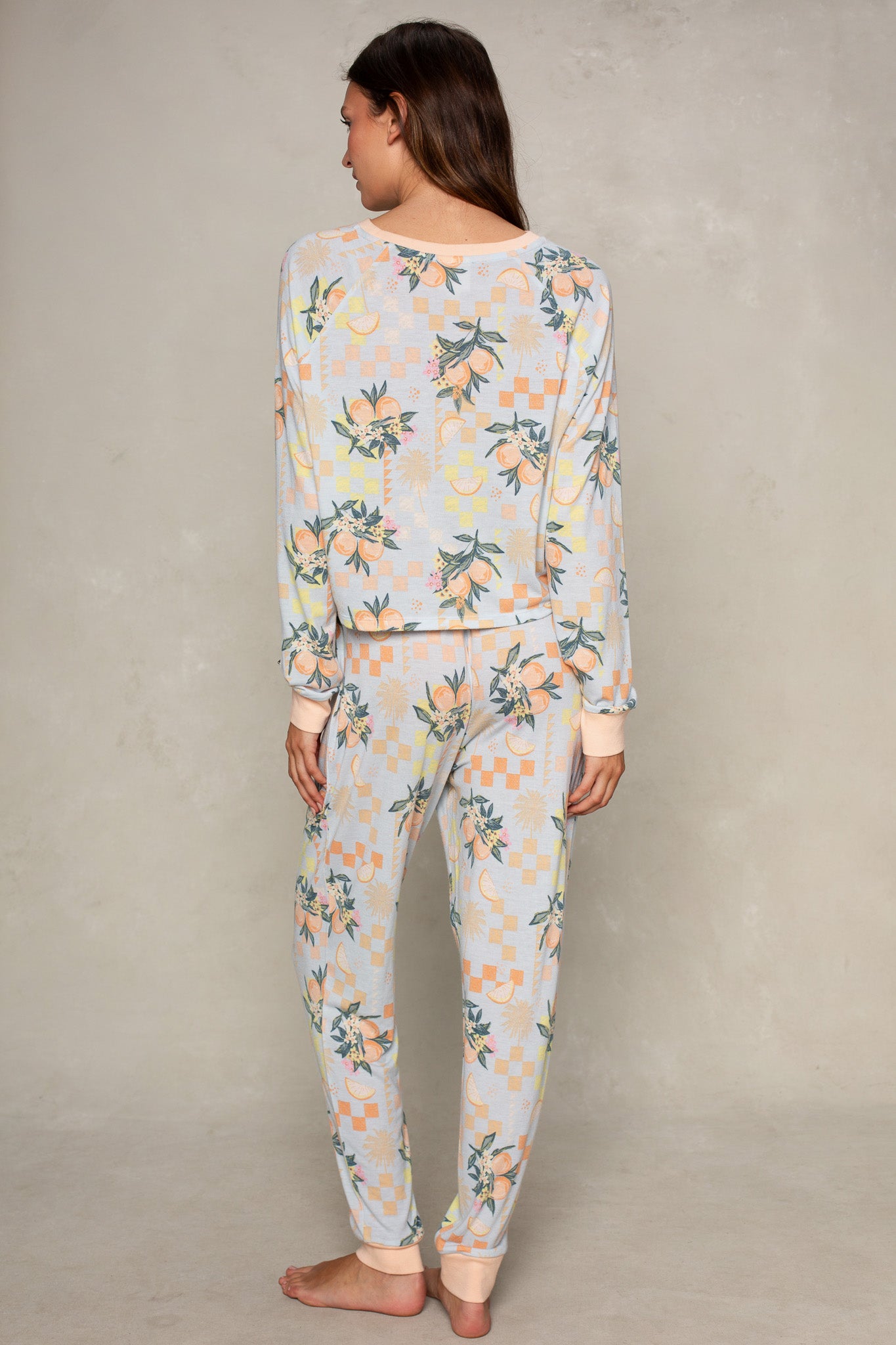 A woman stands barefoot, facing away, wearing Isla's Citrus Check Brushed Jersey Pant—relaxed-fit pastel pajamas with floral and geometric patterns—against a light neutral background.