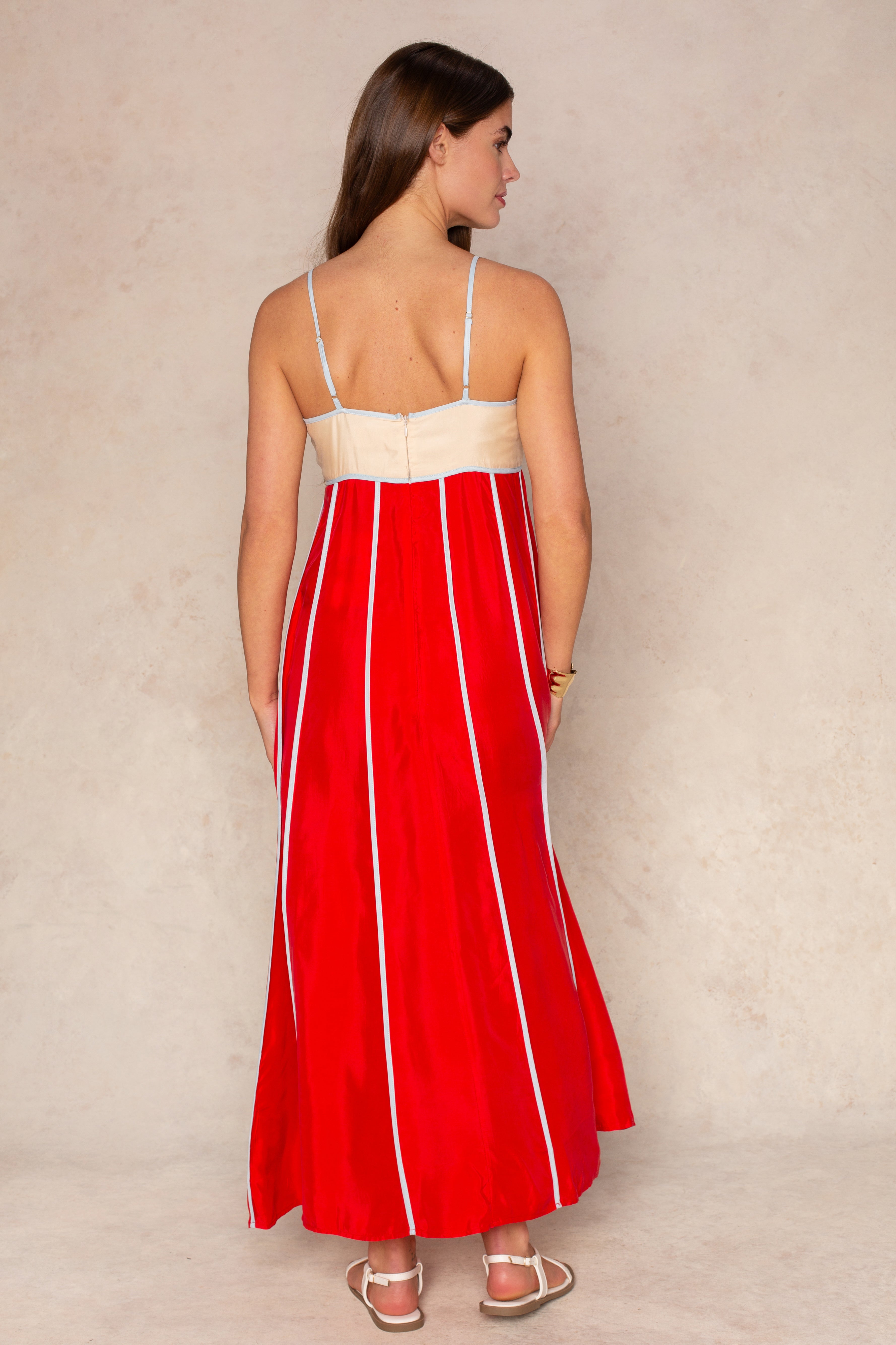 A woman stands with her back to the camera in The Odele Maxi Dress by Sancia—a red, pintucked maxi dress with white vertical stripes and thin straps—paired with white sandals, a bracelet, and loose brown hair against a beige background.