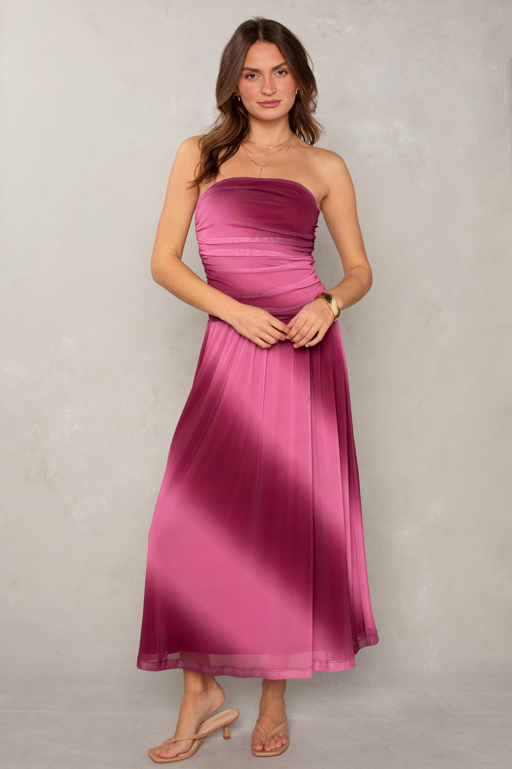 A woman in the Isla Alix Strapless Ruched Mesh Midi Dress stands against a light gray textured wall. She has long brown hair, minimal jewelry, and nude heeled sandals.
