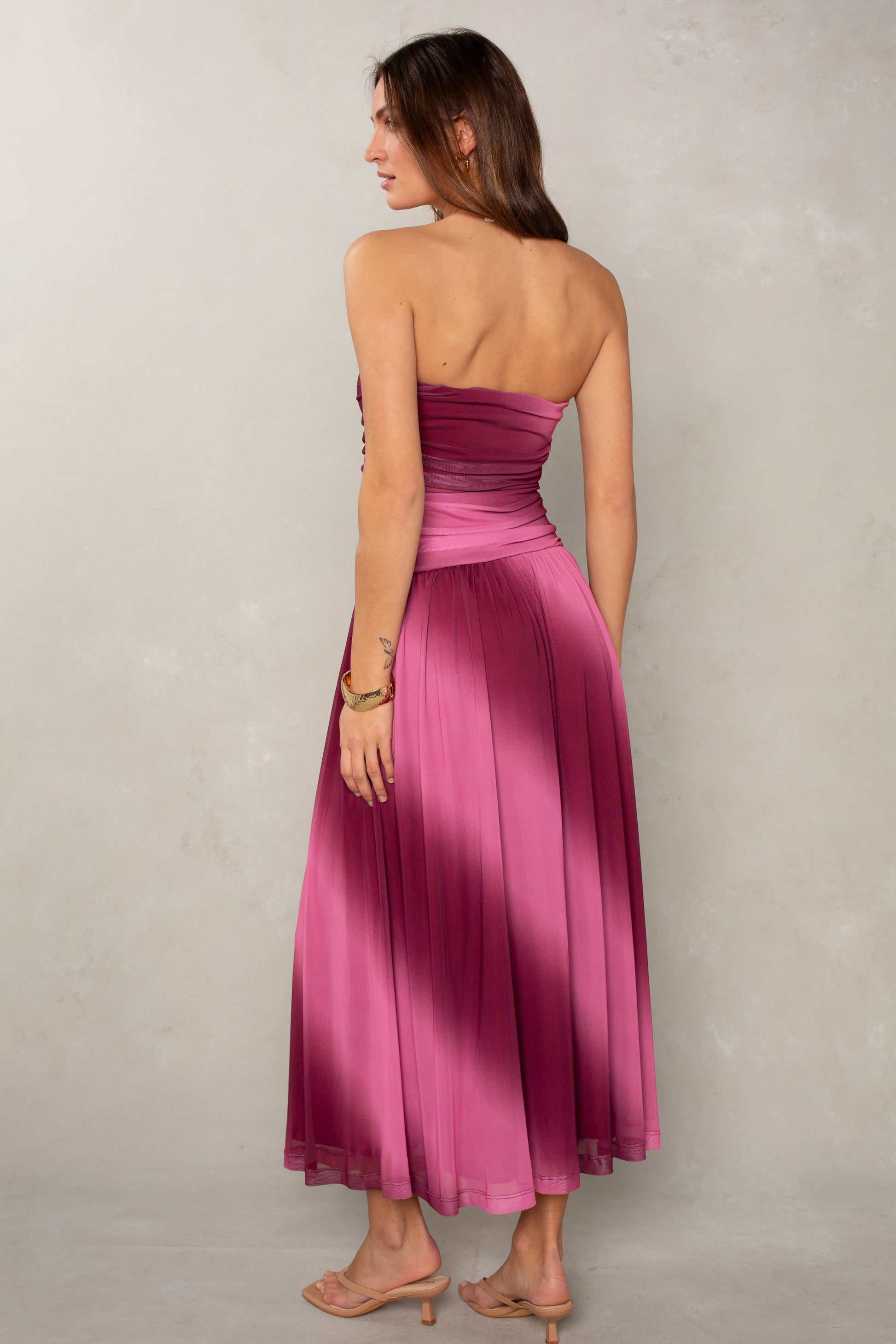 A woman with long brown hair wears the Isla Alix Strapless Ruched Mesh Midi Dress in ombre magenta, featuring a fitted bodice and flowing skirt. She accessorizes with a bracelet and beige heeled sandals against a light gray textured wall.