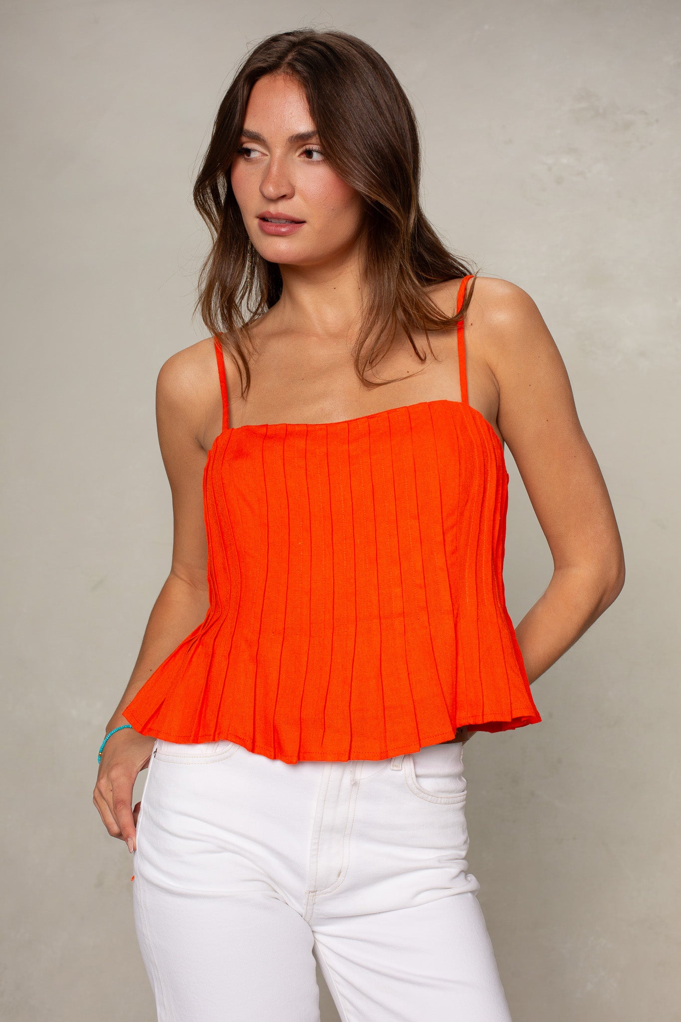 A woman stands against a light gray background, looking slightly to her left with a relaxed expression, wearing the Isla Margot Top in bright orange—crafted from premium materials—and white pants.