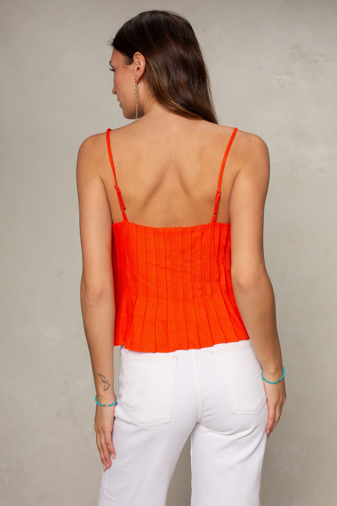 A woman with long brown hair wears the Isla Margot Top—a bright orange, sleeveless, pleated style with thin straps—paired with high-waisted white pants. She accessorizes with dangle earrings and blue beaded bracelets against a neutral background.