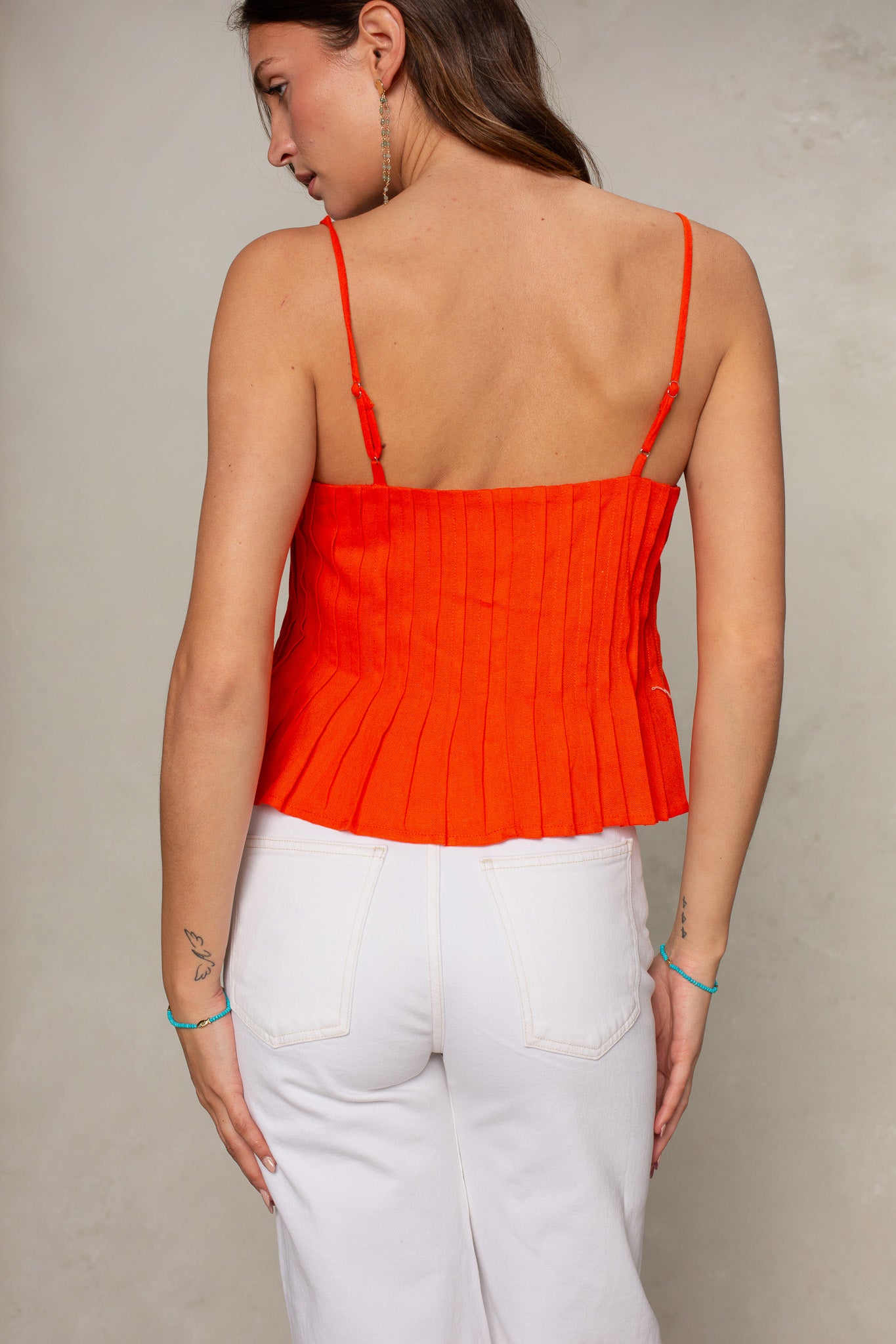 A woman faces away from the camera, wearing the Isla Margot Top in bright orange with white high-waisted pants. Her hair is down and small tattoos are visible on her arms.