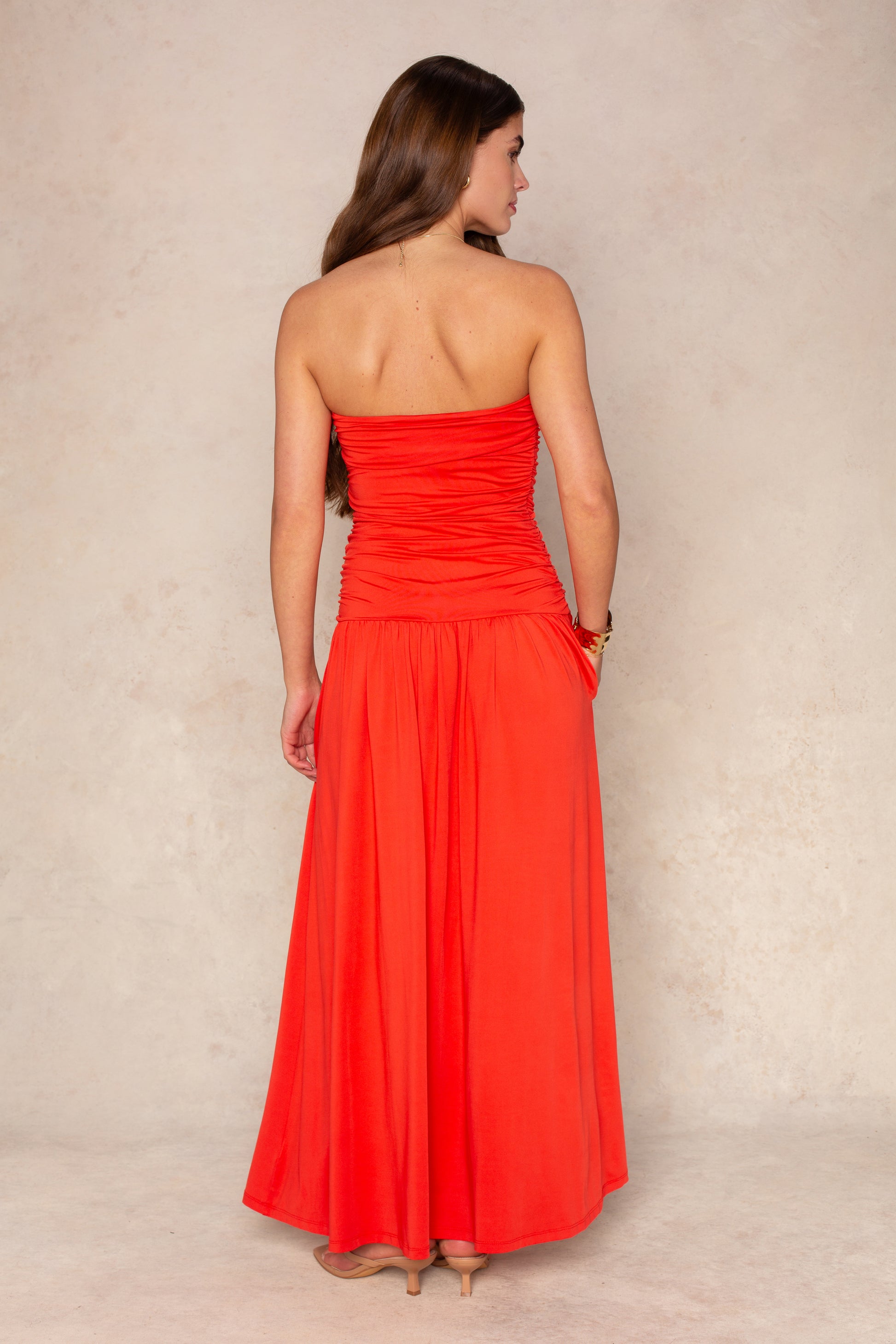 A woman with long brown hair, facing away, wears the Nia Santal Midi Dress—a strapless bright red-orange ruched-bodice dress flowing to her ankles—with beige heels, set against a neutral textured background.