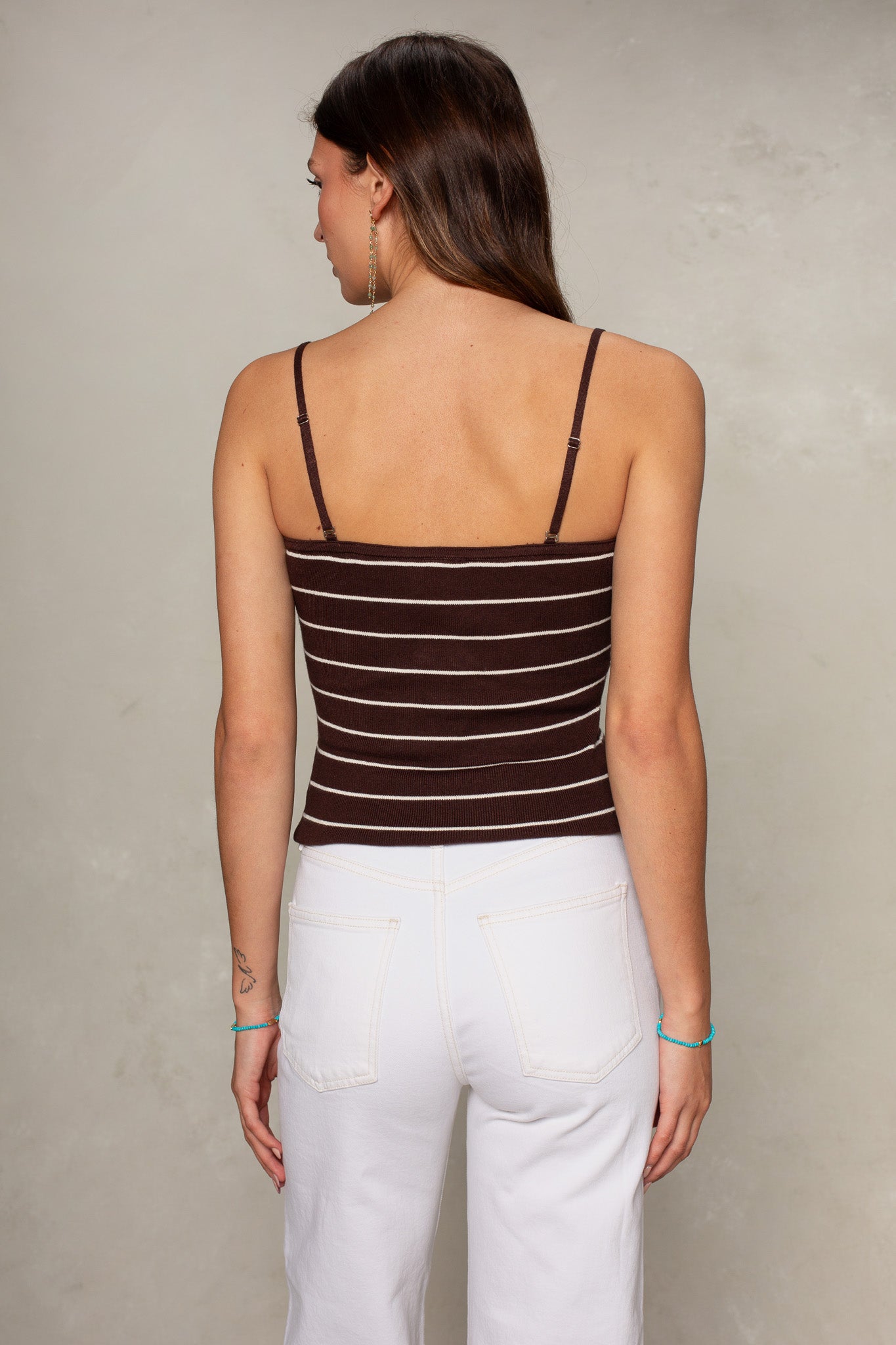 A woman with long brown hair stands facing away, wearing the Isla Chelsea Convertible Cami with adjustable straps and high-waisted white jeans. She has a small tattoo on her left arm and a blue bracelet on her right wrist.