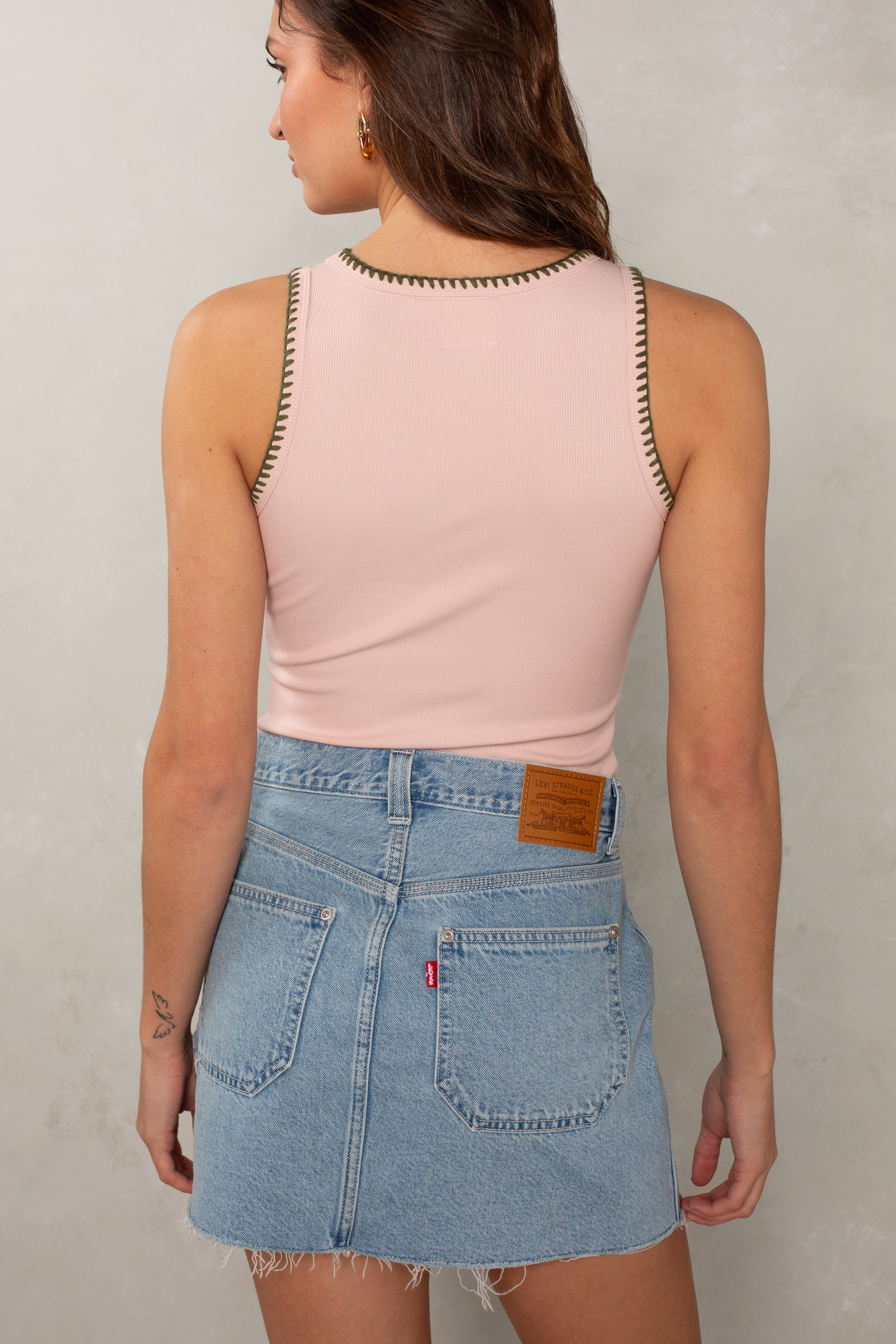 A woman faces away, wearing the Isla Simone Stitch Tank in light pink with dark trim, paired with a high-waisted light blue denim mini skirt. Visible pockets, detailed stitching, wavy brown hair, and a small arm tattoo complete the look.