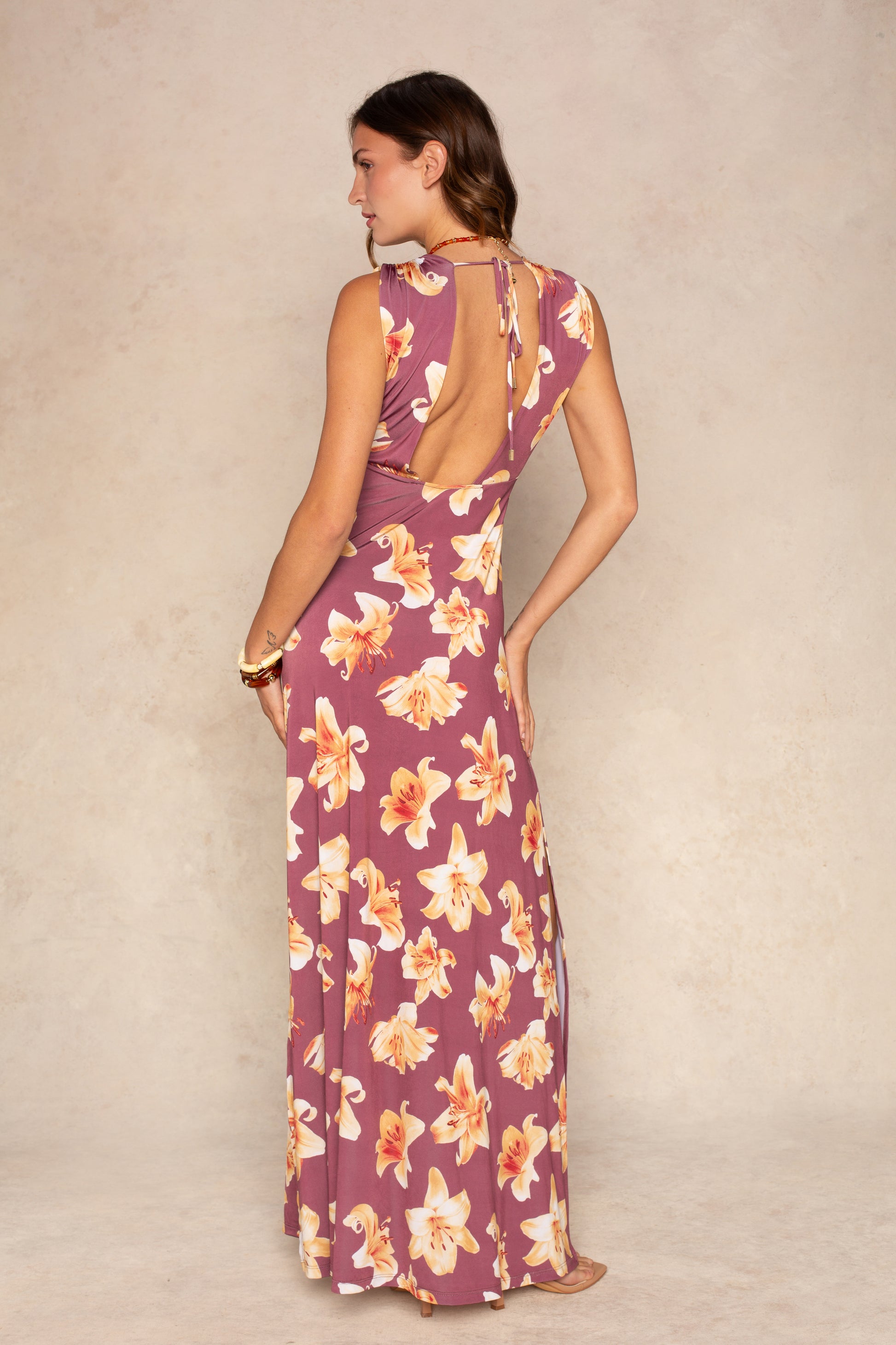 A woman poses barefoot against a neutral backdrop wearing the Isla Raven Plunging Maxi Dress, a sleeveless mauve summer dress featuring yellow and white floral print, a plunging open back, and charming ruffled details.