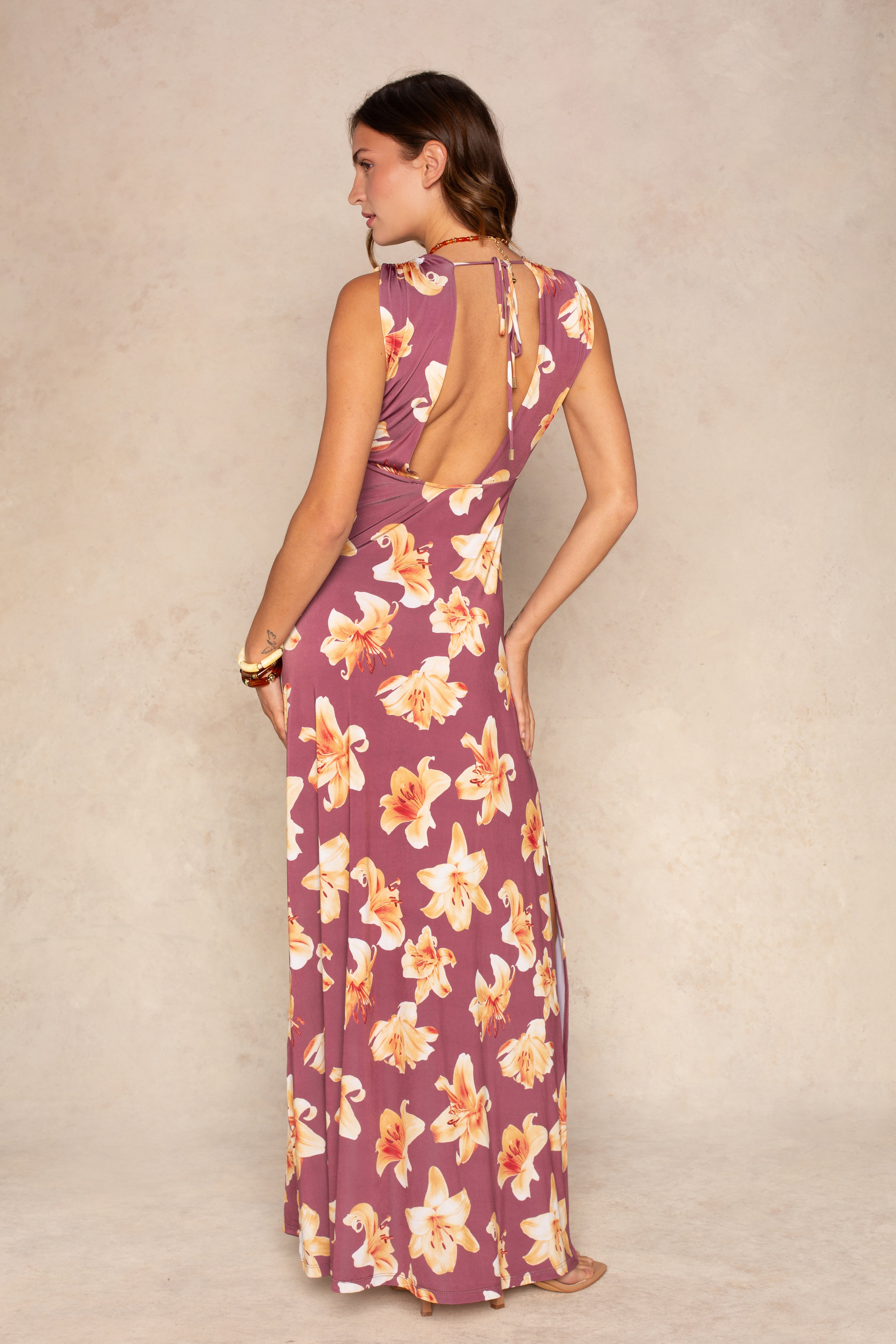 A woman poses barefoot against a neutral backdrop wearing the Isla Raven Plunging Maxi Dress, a sleeveless mauve summer dress featuring yellow and white floral print, a plunging open back, and charming ruffled details.