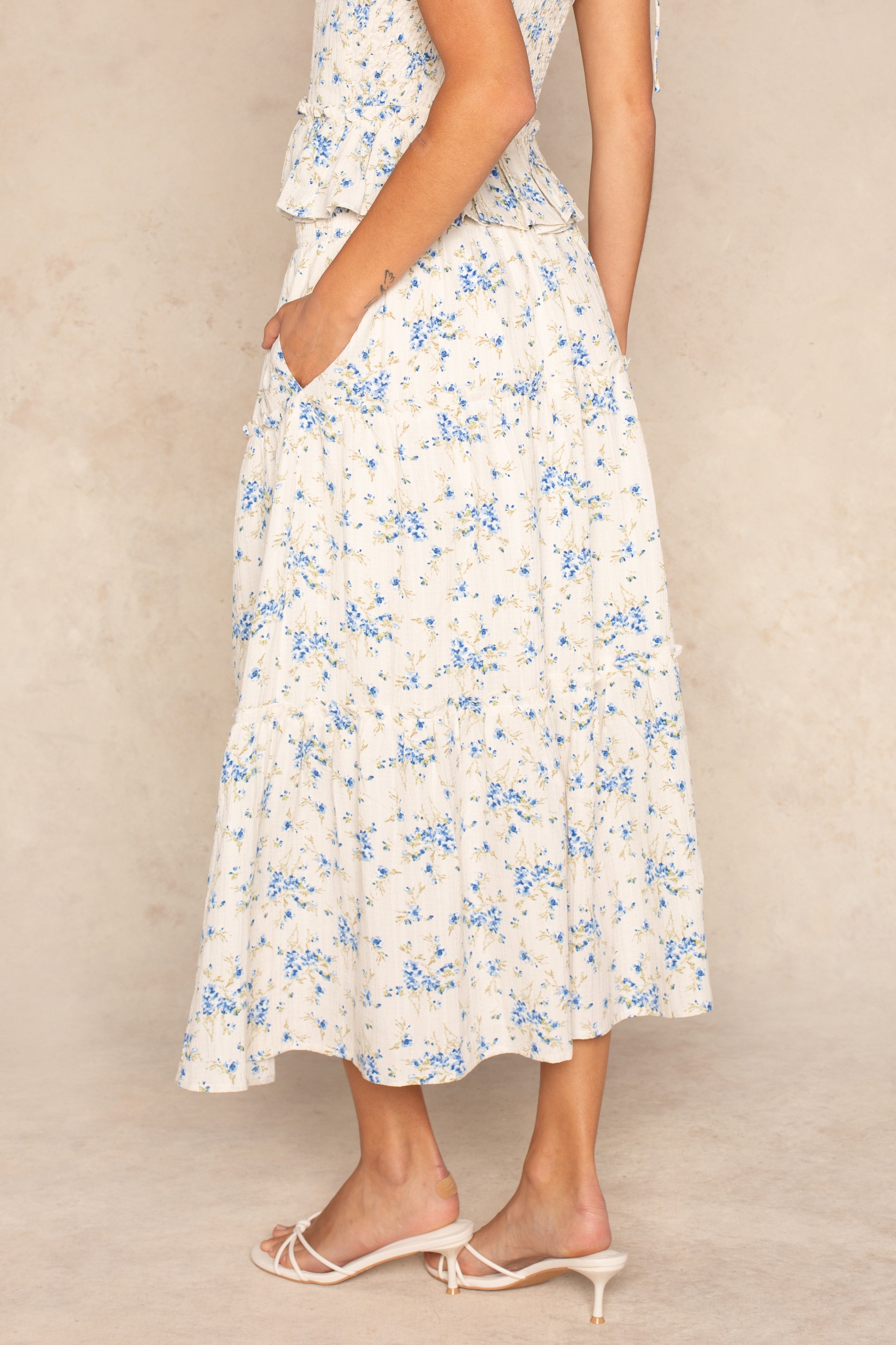 A woman stands sideways in the Isla Edie Maxi Floral Tiered Skirt, featuring an off-white base with blue floral print and a comfy elastic waistband. She pairs it with a matching top and white low-heeled sandals against a beige background.