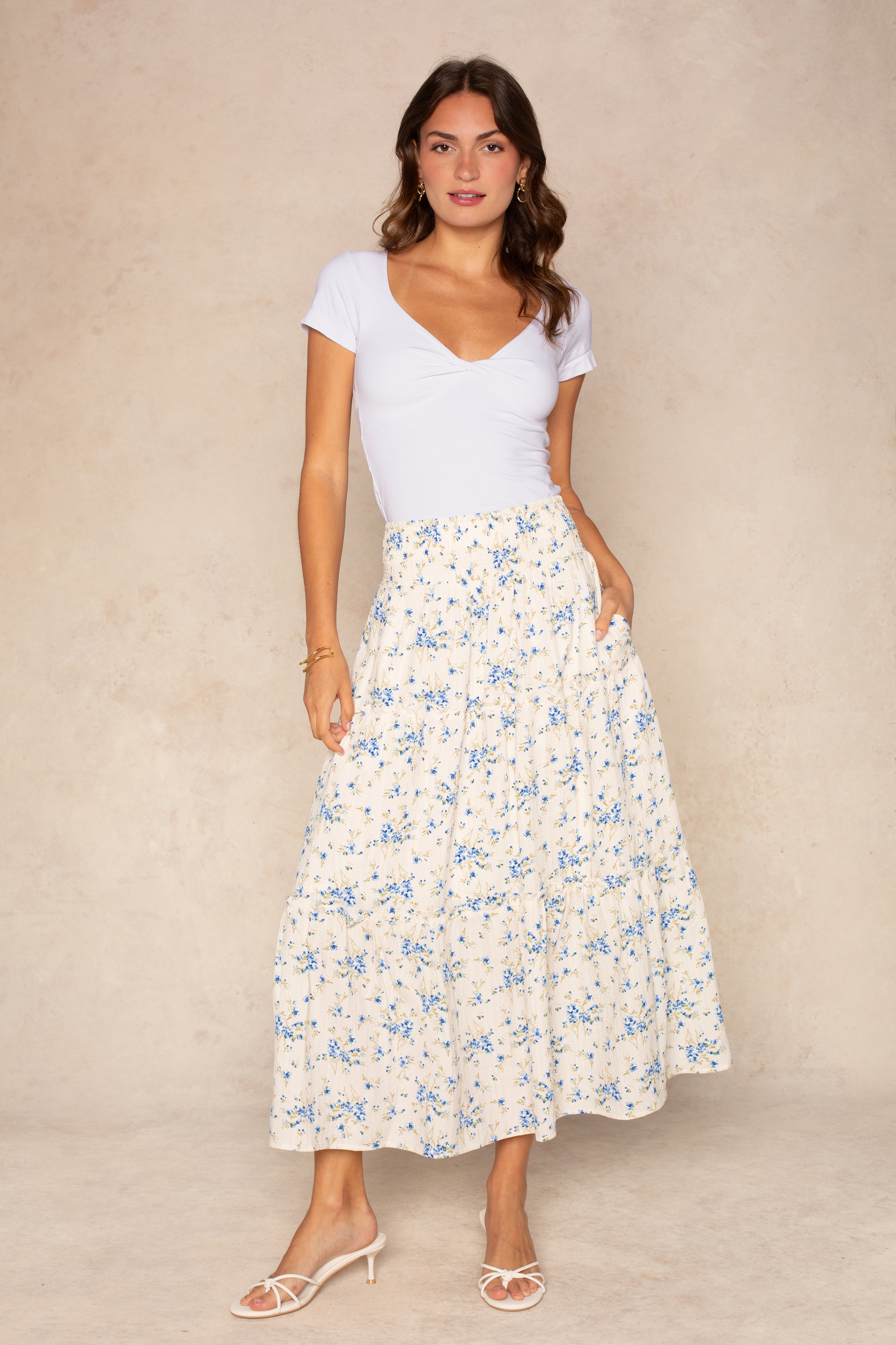 A woman stands against a beige background wearing the Isla Edie Maxi Floral Tiered Skirt in White Multi with a fitted white short-sleeve top, white heeled sandals, and gold jewelry.