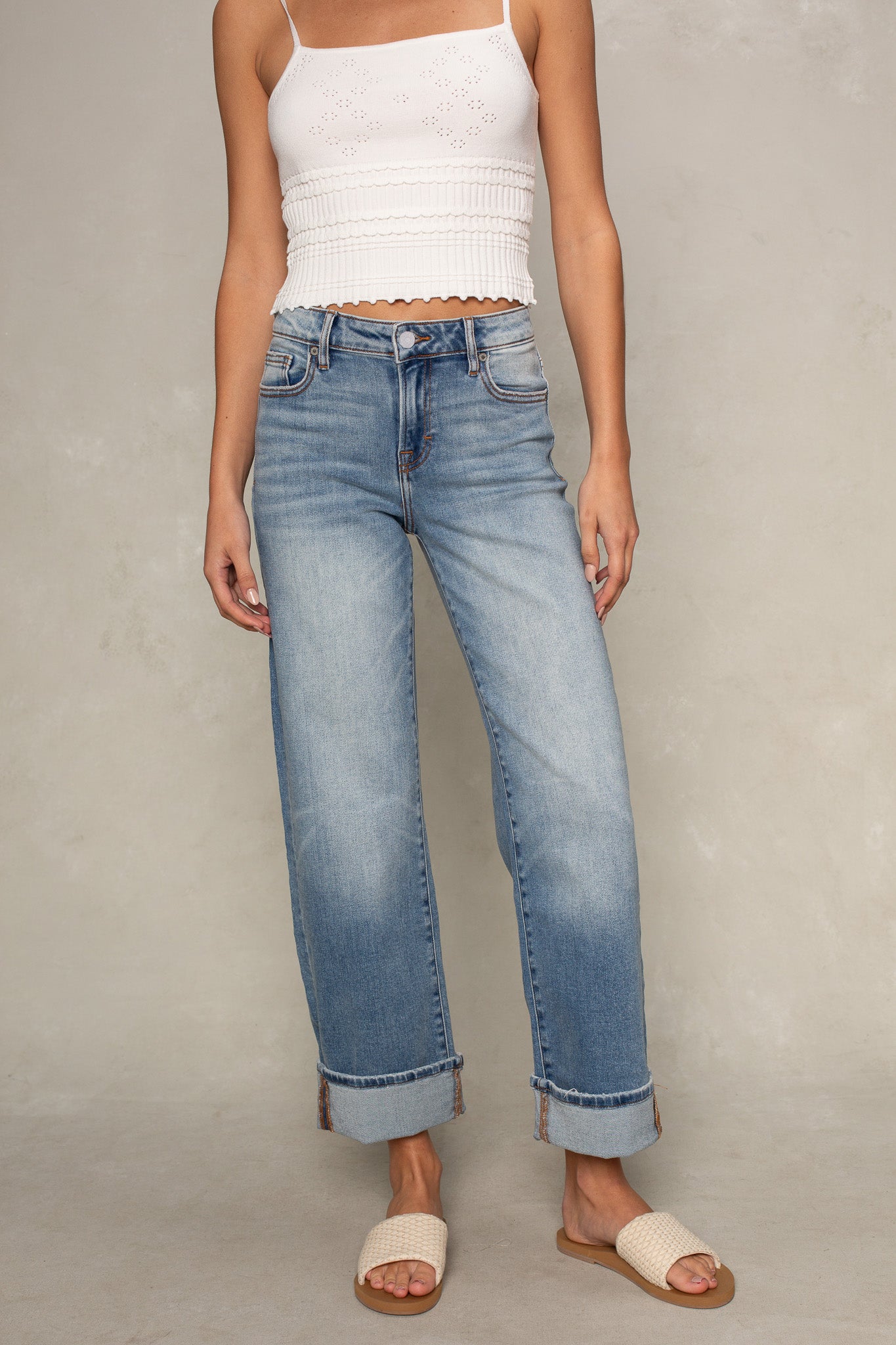 A woman wears a white sleeveless crop top, Isla Dylan Mid Rise Cuffed Dad Jean in medium denim, and beige woven slide sandals against a neutral background. Her head is not visible in the image.