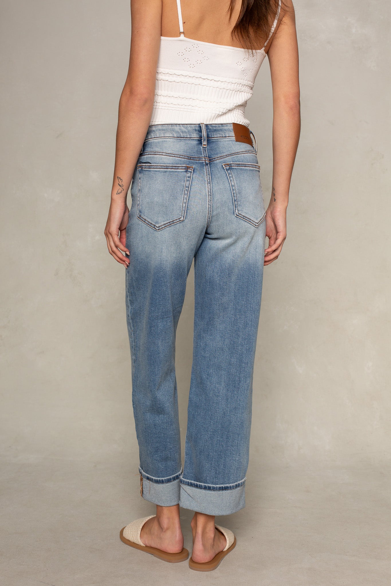 A person stands facing away against a plain light background, wearing Isla's Dylan Mid Rise Cuffed Dad Jean in light blue, paired with a white sleeveless top and woven slip-on sandals.