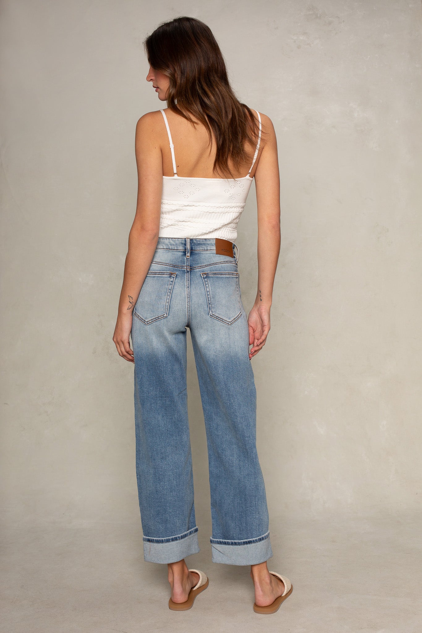 A woman faces away, wearing the Isla Quinn Pointelle Cami and light blue high-waisted wide-leg jeans with cuffed hems, paired with tan sandals. The background is a plain, light-colored wall.