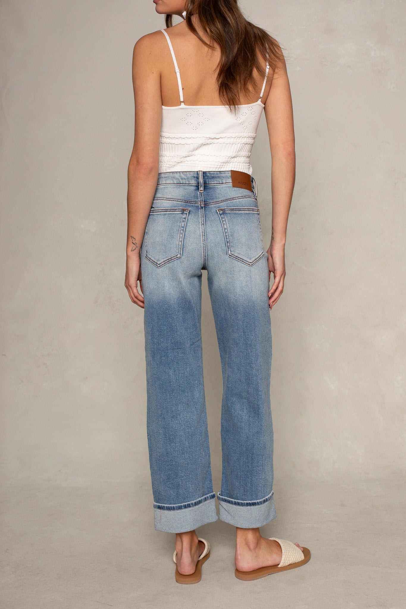 A woman faces away wearing a white tank top, Isla Dylan Mid Rise Cuffed Dad Jean, and woven sandals. She stands in front of a plain light-colored wall.