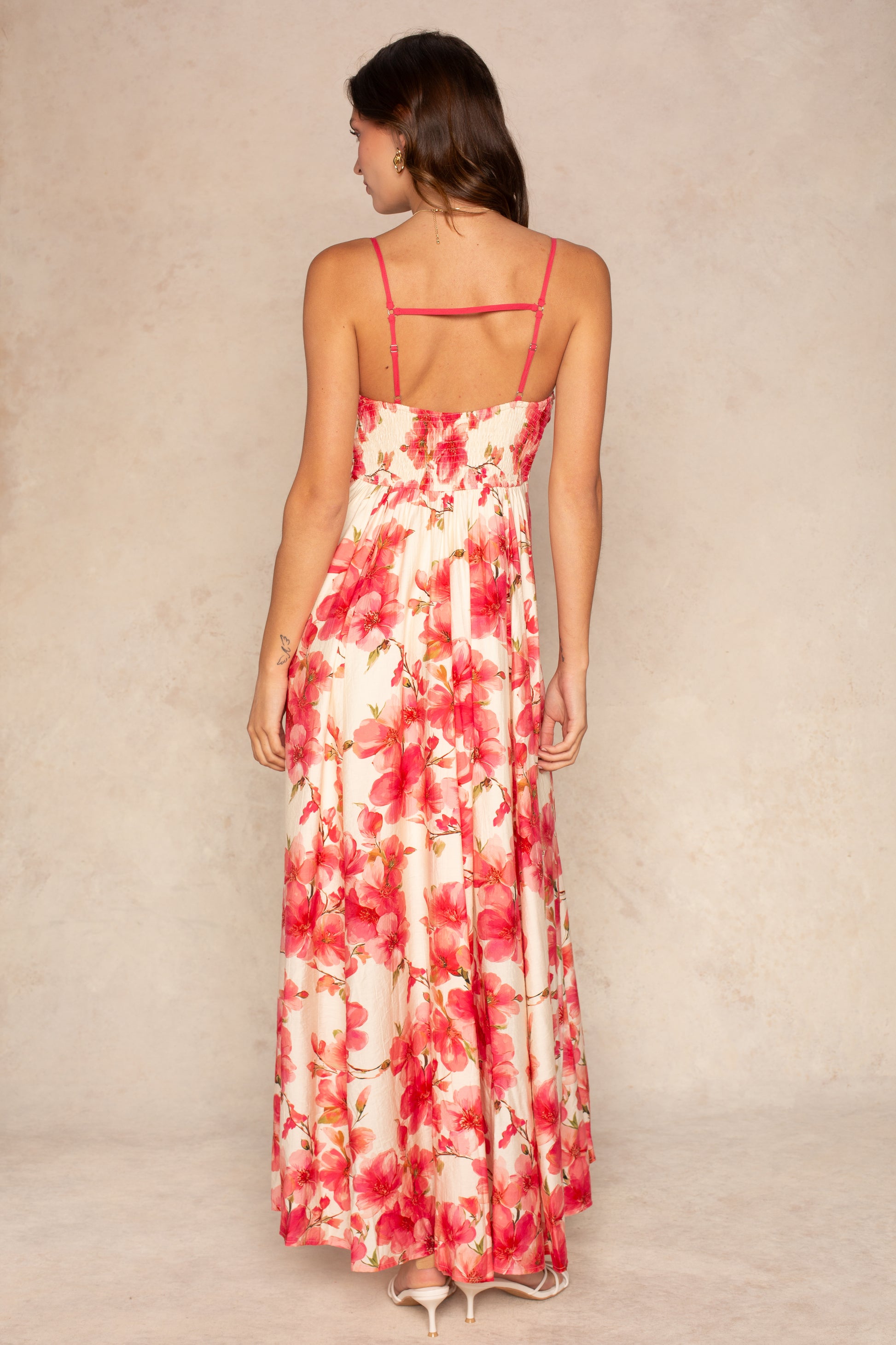 A woman with medium-length brown hair faces away from the camera, modeling the Isla Dannah Maxi Dress—a long, flowy vacation dress with adjustable spaghetti straps, an open back, and vibrant pink and red tropical floral patterns.