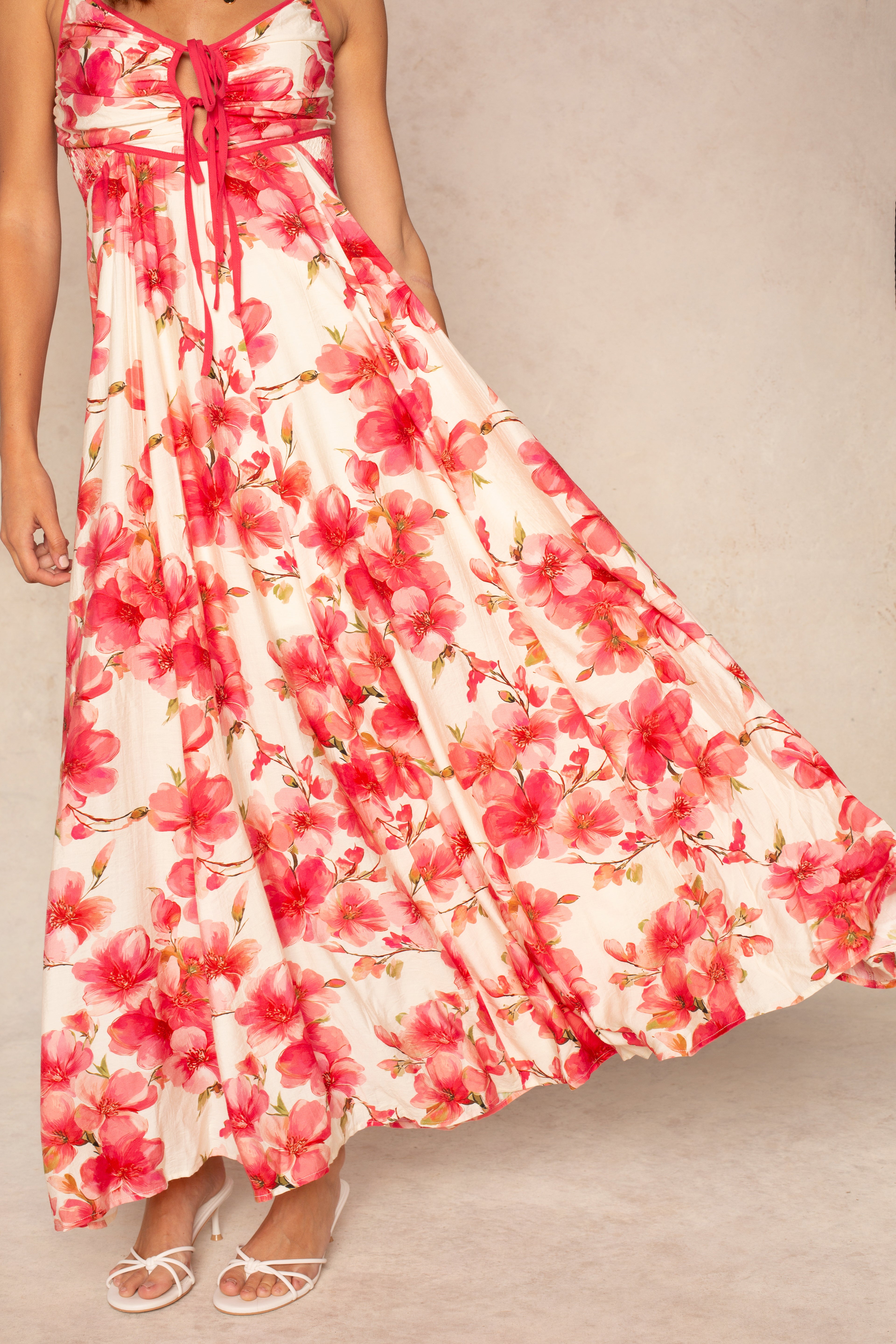Someone models the Isla Dannah Maxi Dress, featuring a tropical floral print, adjustable spaghetti straps, and a tie-front bodice with white strappy heels against a light beige background—ideal for your next vacation look.