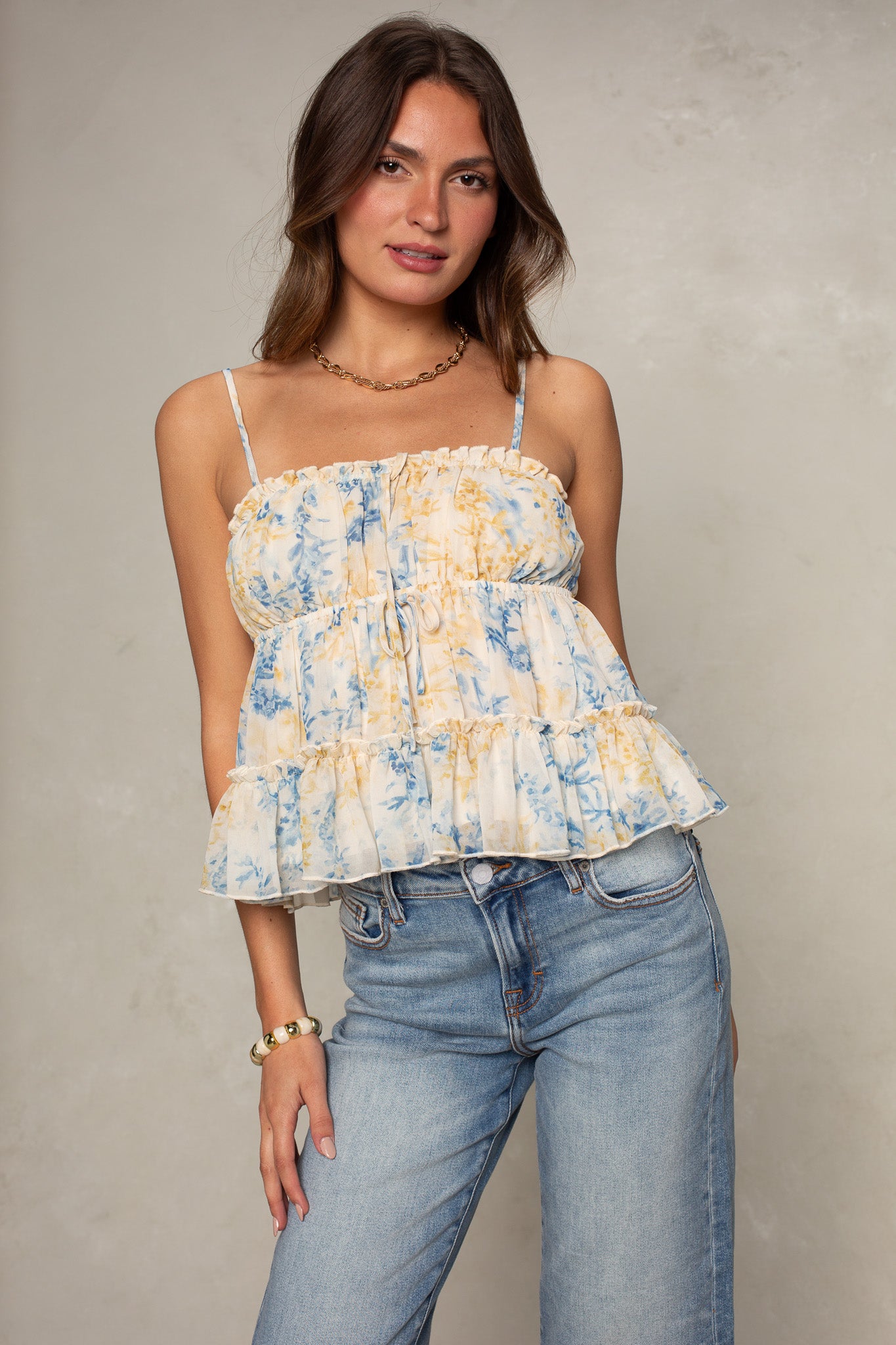A woman showcases Isla’s Tiered Chiffon Cami in Blue Multi—featuring ruffles and a floral print—paired with light blue jeans, gold jewelry, and loose brown hair, set against a neutral background for an effortlessly chic look.