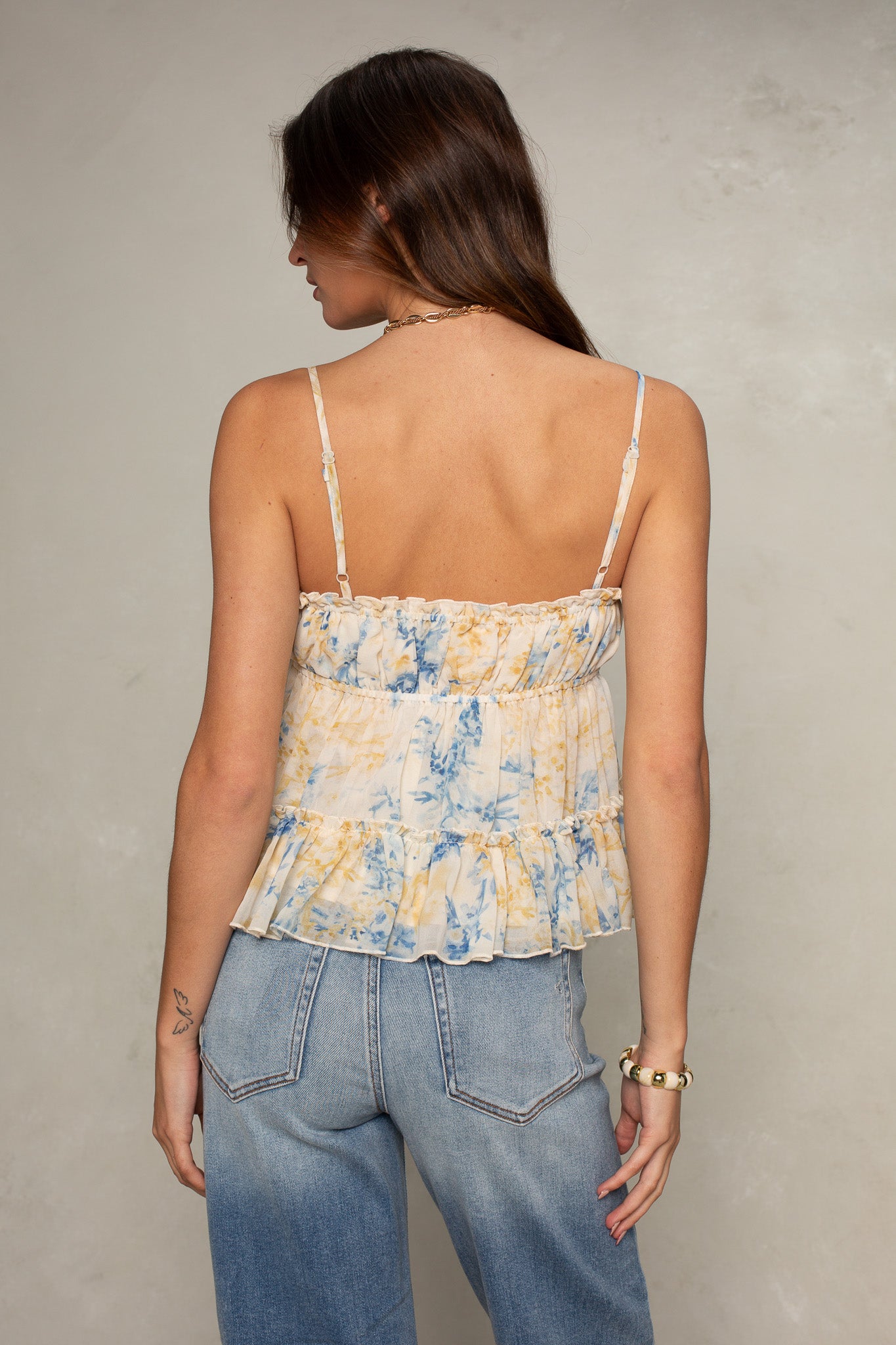 A woman with long brown hair is seen from the back wearing Isla's Tiered Chiffon Cami, a sleeveless tie-dye blue and yellow top, paired with high-waisted blue jeans against a neutral light background.