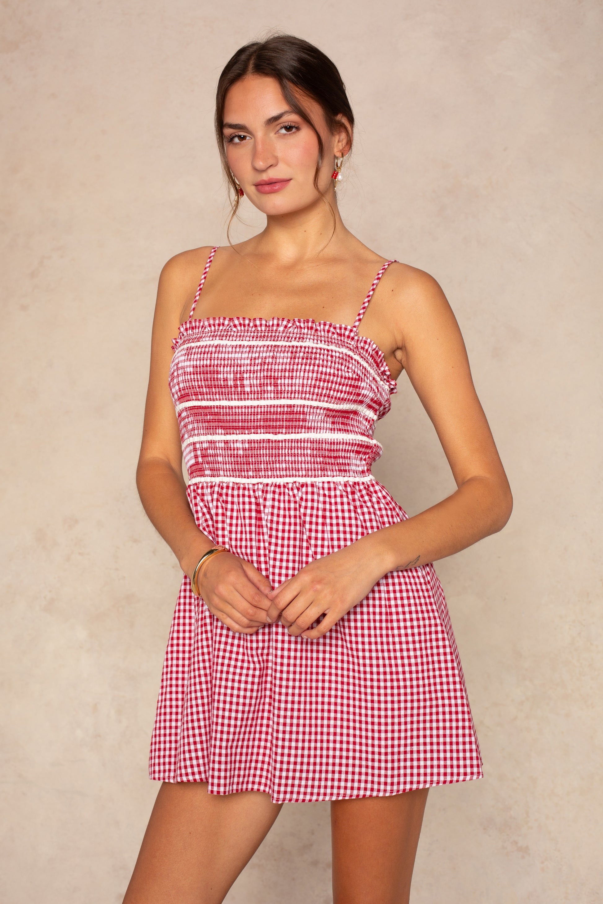 A woman wears the Isla Jillian Smocked Fit And Flare Mini Dress in Red Multi, featuring a sleeveless design, thin straps, and a smocked bodice, standing against a neutral background and smiling slightly at the camera.
