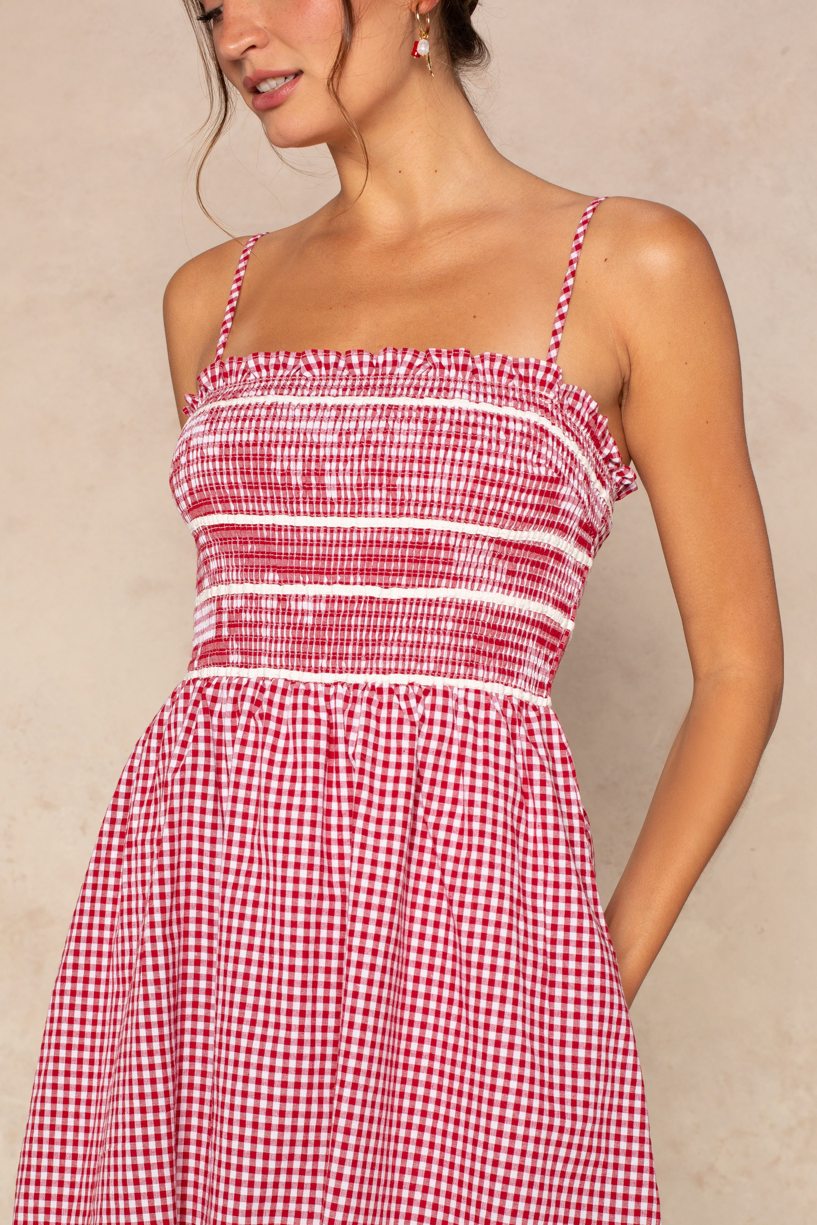 A woman wears the Isla Jillian Smocked Fit And Flare Mini Dress, featuring red gingham fabric, thin straps, ruffle details, and a smocked bodice. She stands with one hand in her pocket against a neutral background; her face is partly out of frame.