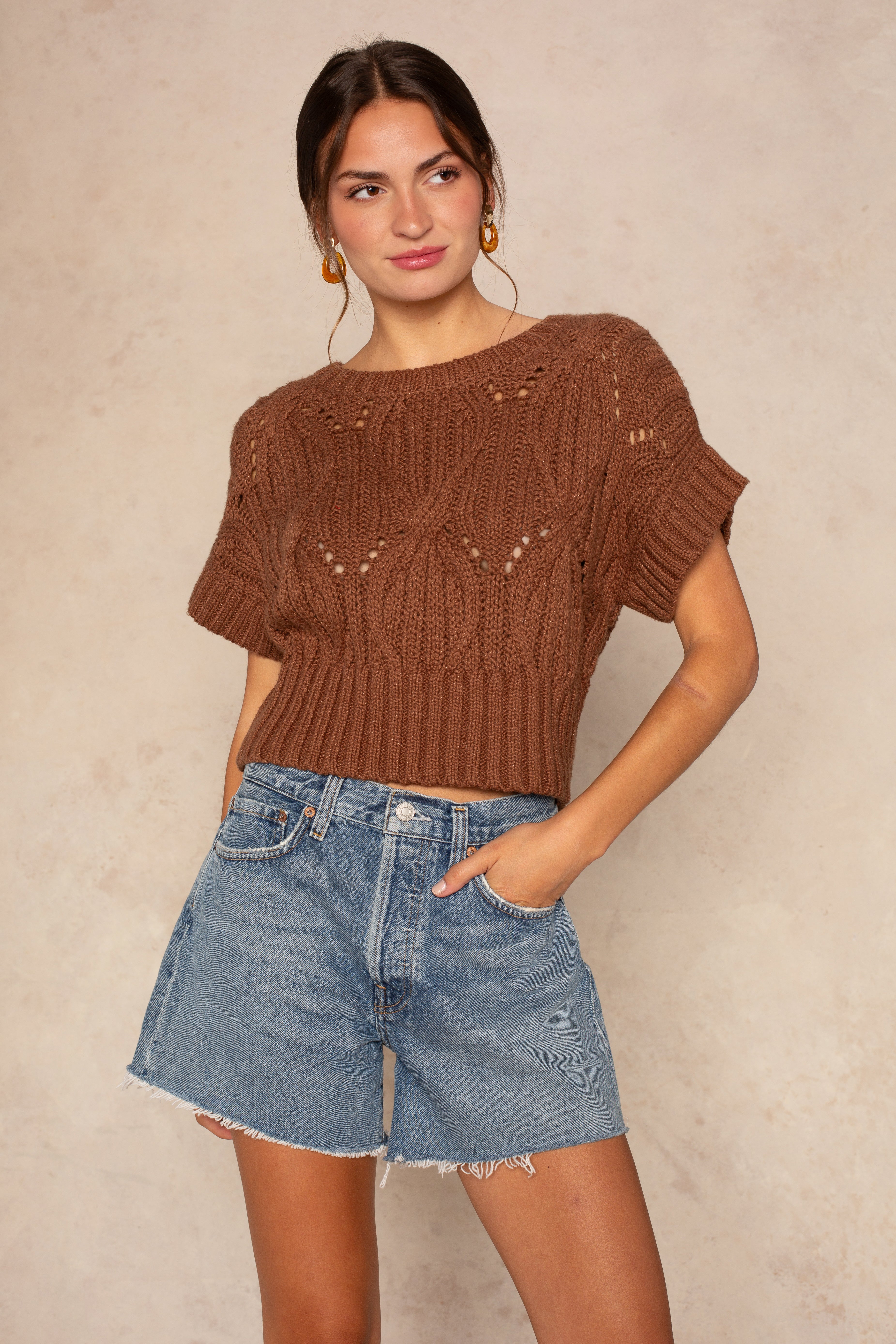 A woman stands against a beige background wearing the Isla Cali Crochet sweater in dark brown and high-waisted light blue denim shorts with frayed hems, one hand in her pocket as she looks slightly to the side.