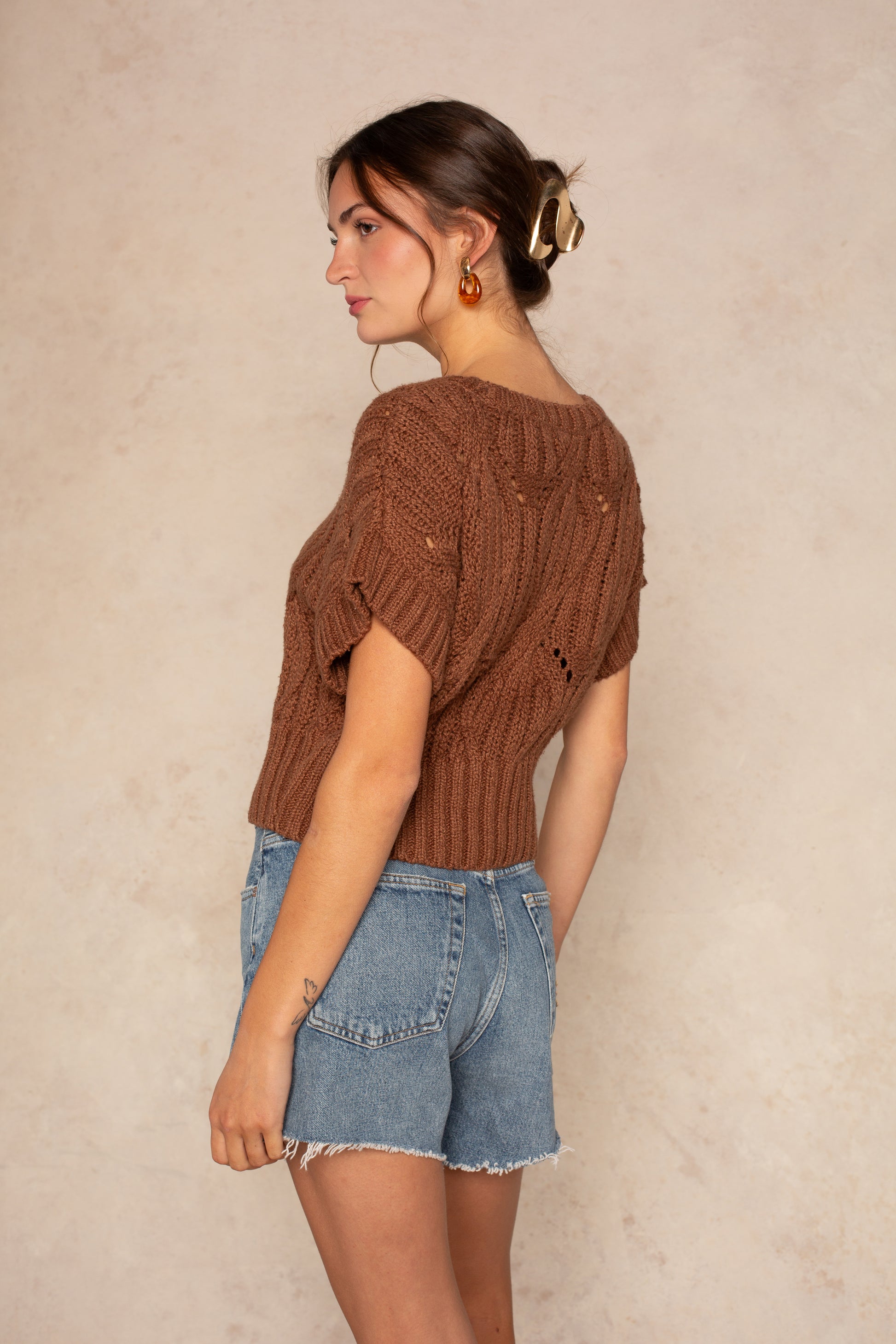 A woman stands sideways against a beige background, wearing Isla's Cali Crochet short-sleeve top in brown, light blue denim shorts, gold earrings, and a hair clip holding her brown hair in a low bun.
