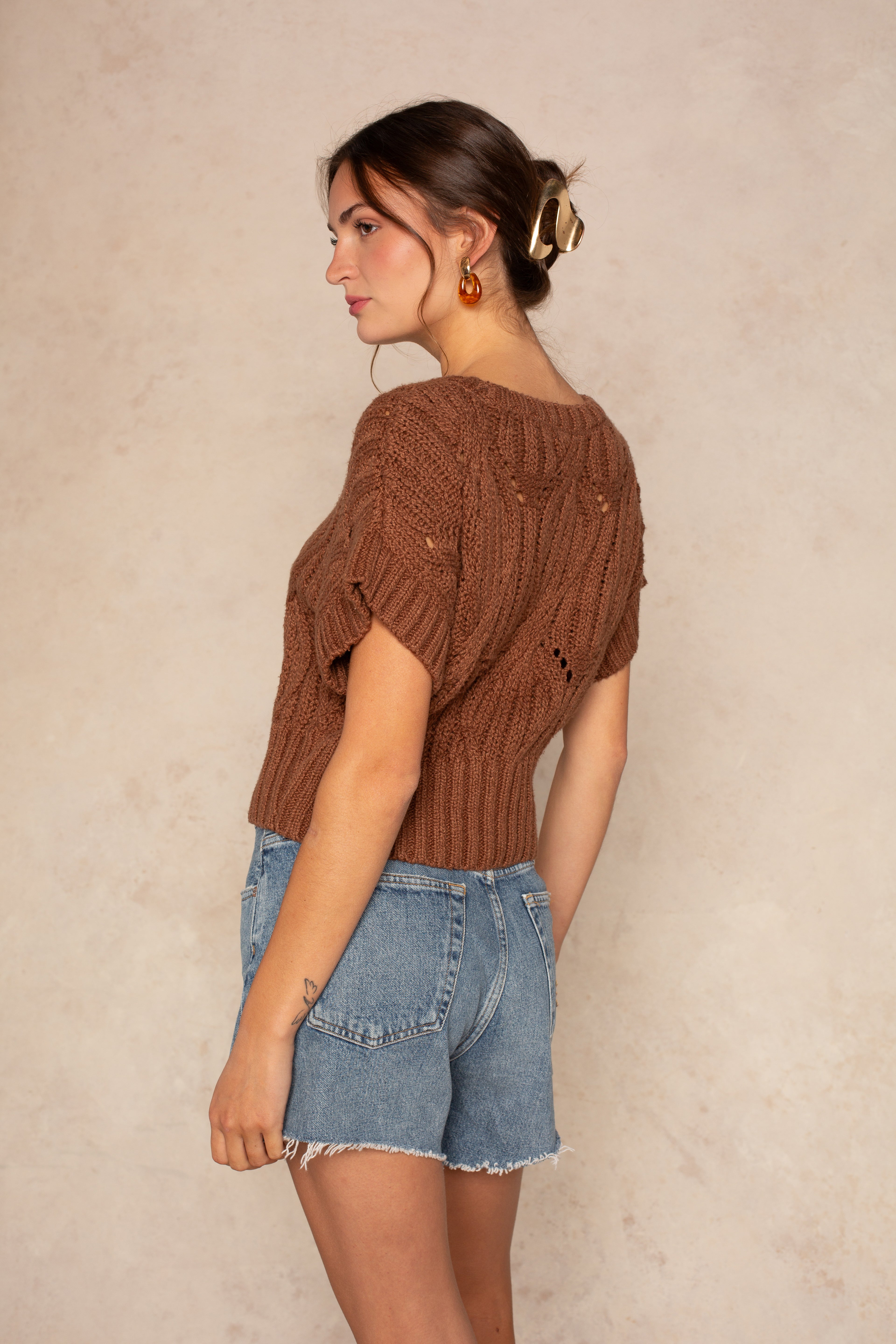A woman stands sideways against a beige background, wearing Isla's Cali Crochet short-sleeve top in brown, light blue denim shorts, gold earrings, and a hair clip holding her brown hair in a low bun.